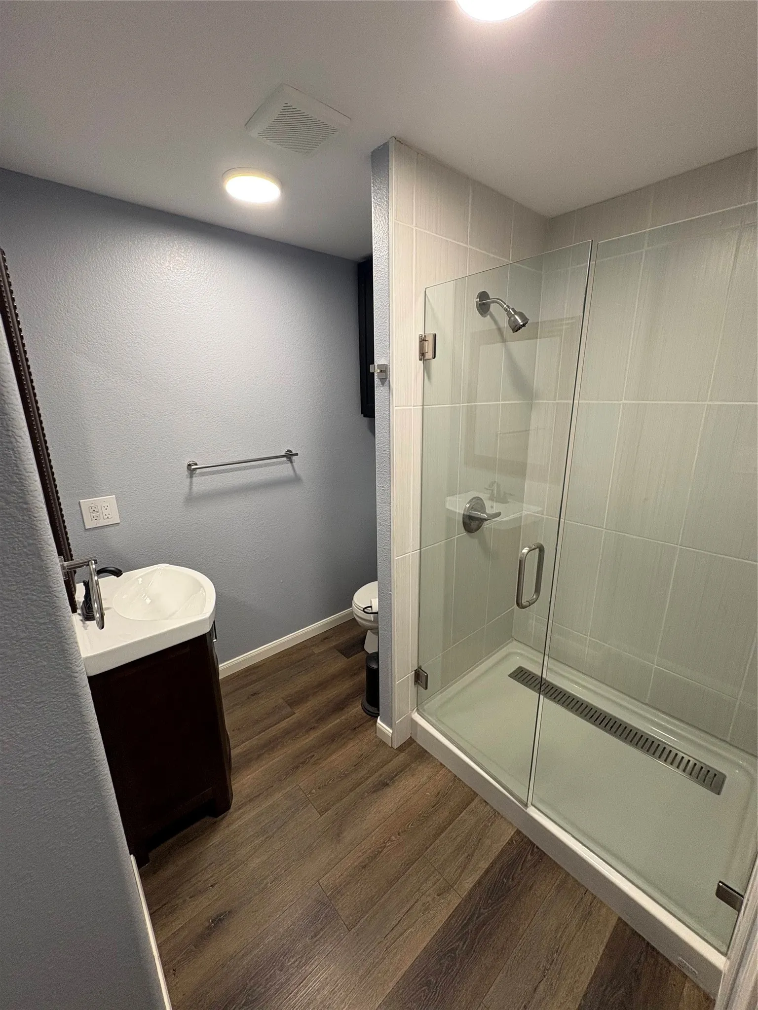 Glass-enclosed shower with a linear drain, large format tile shower surround, wood-finish flooring, and a single-sink vanity with a vessel sink