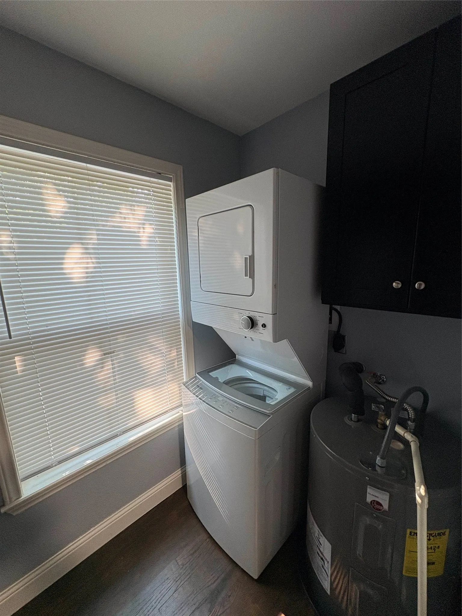 Stackable washer and dryer unit, dark wood-finish flooring, window with blinds, upper cabinetry, and water heater