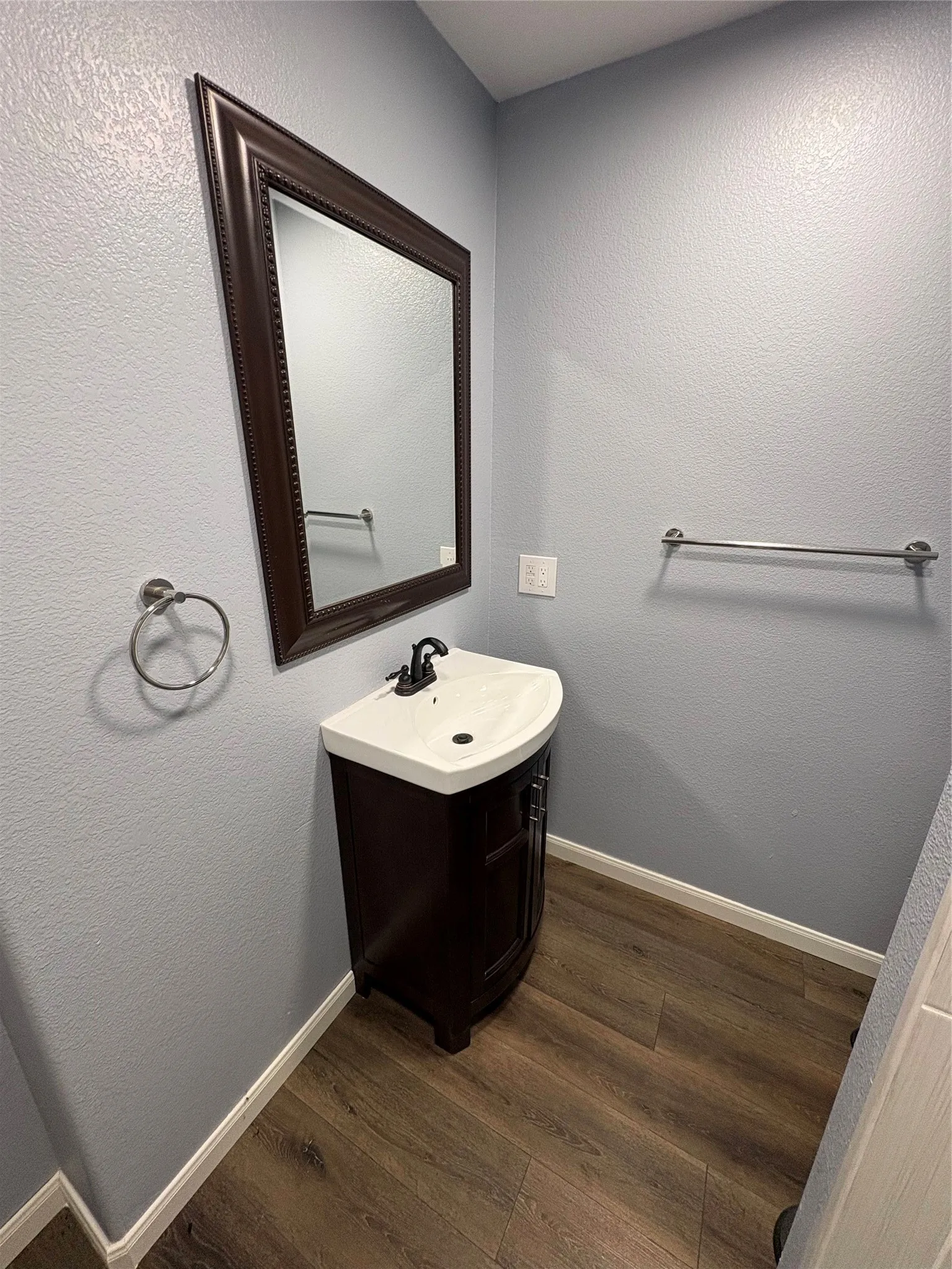 Compact powder room featuring a sleek pedestal sink with single-handle faucet, wood-finish vanity cabinet, framed mirror with dark wood trim, chrome towel bar and ring hardware, and wood-finish flooring throughout