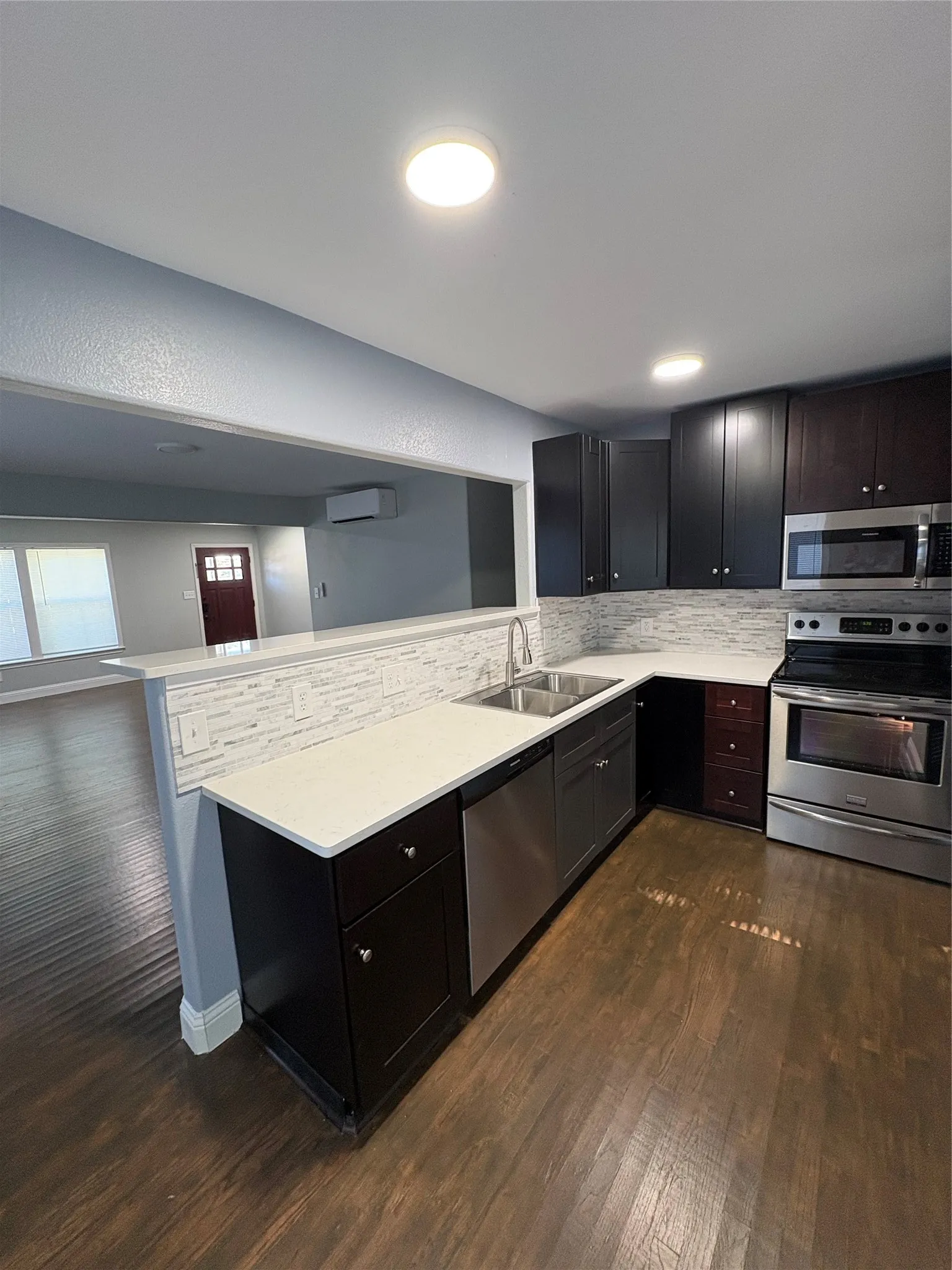 Modern kitchen featuring a large center island with white quartz countertop and undermount sink, black cabinetry with hardware accents, stainless steel appliances including double wall ovens, subway tile backsplash, and warm wood-finish flooring throughout the open-concept space
