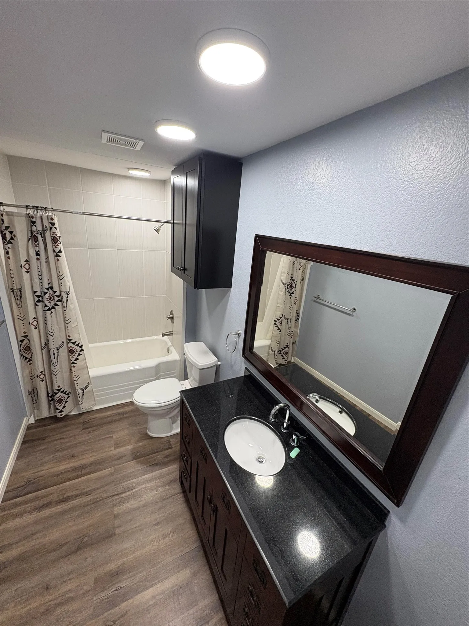 Wood-finish flooring, a white bathtub with tile surround, and a dark wood vanity with a dark countertop and integrated sink