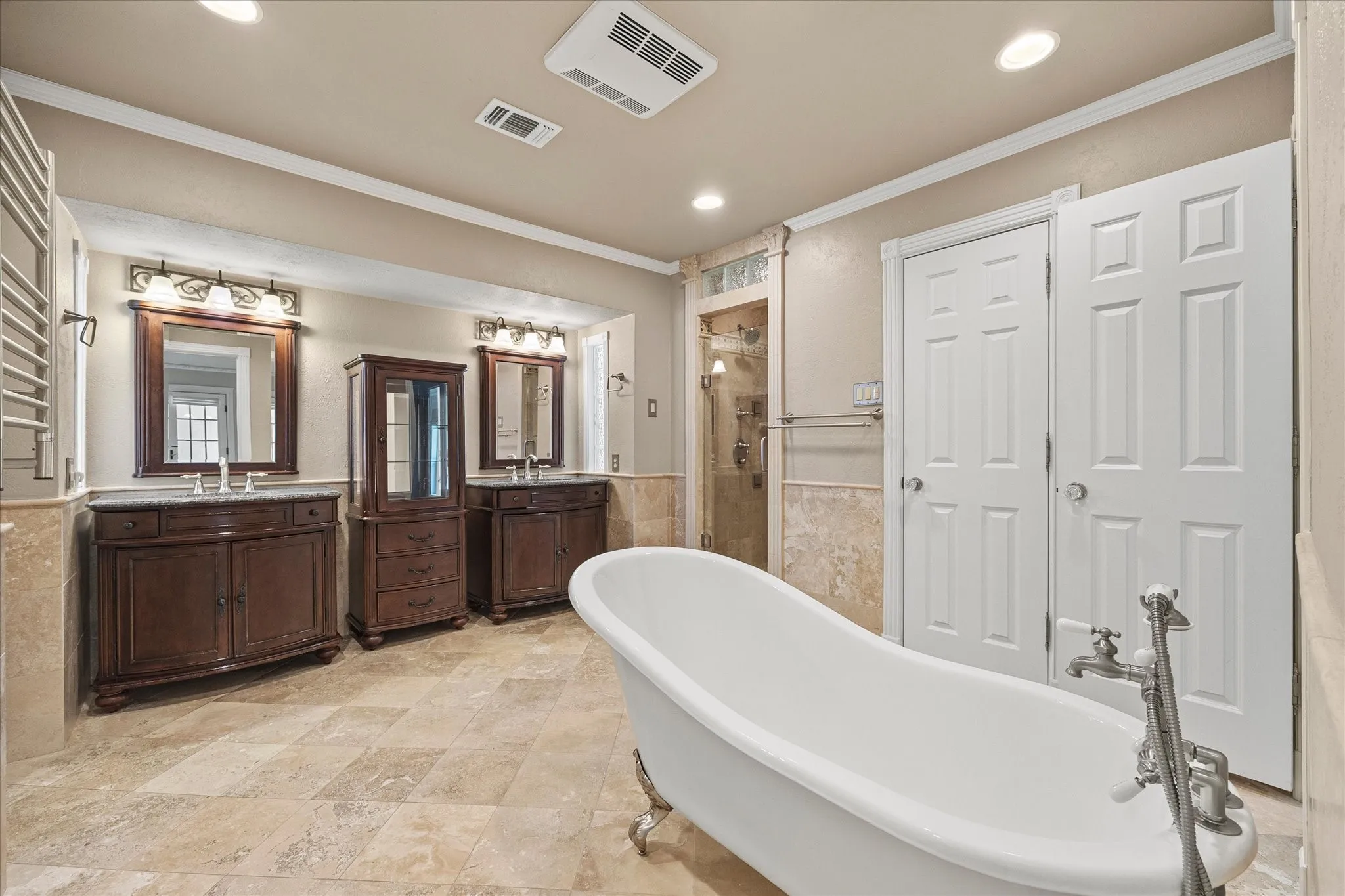 Update primary bath featuring a freestanding slipper tub, dual vanities, travertine flooring, and walk-in shower.  This spa-type retreat also features two closets on each side.
