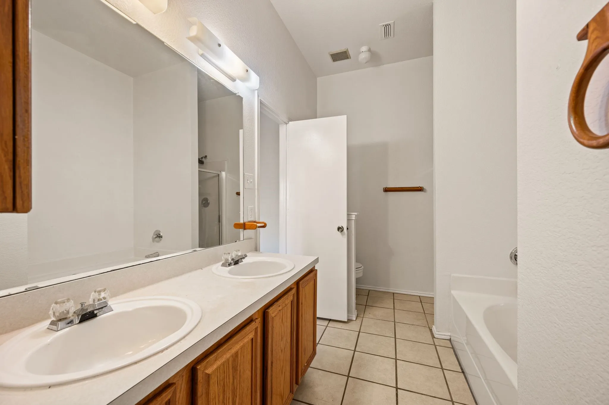 Dual basin vanity with wood-finish cabinetry, a full-width mirror, tile flooring, and a built-in bathtub