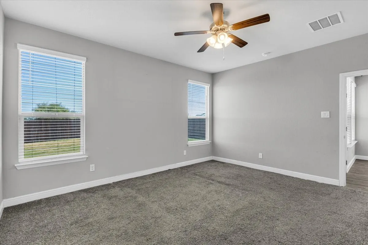 Neutral gray walls, plush gray carpeting, two windows with white blinds, and a ceiling fan with light fixture