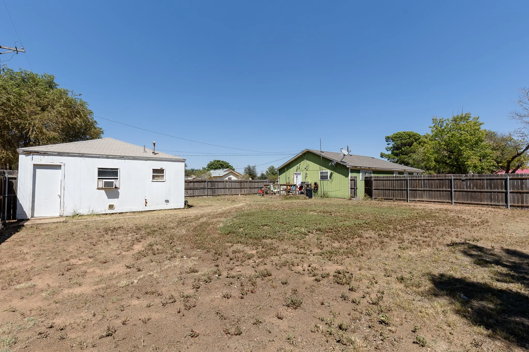 Spacious lot with expansive grounds featuring mature shade trees, wooden perimeter fencing, and multiple outbuildings including a white storage structure and green shed