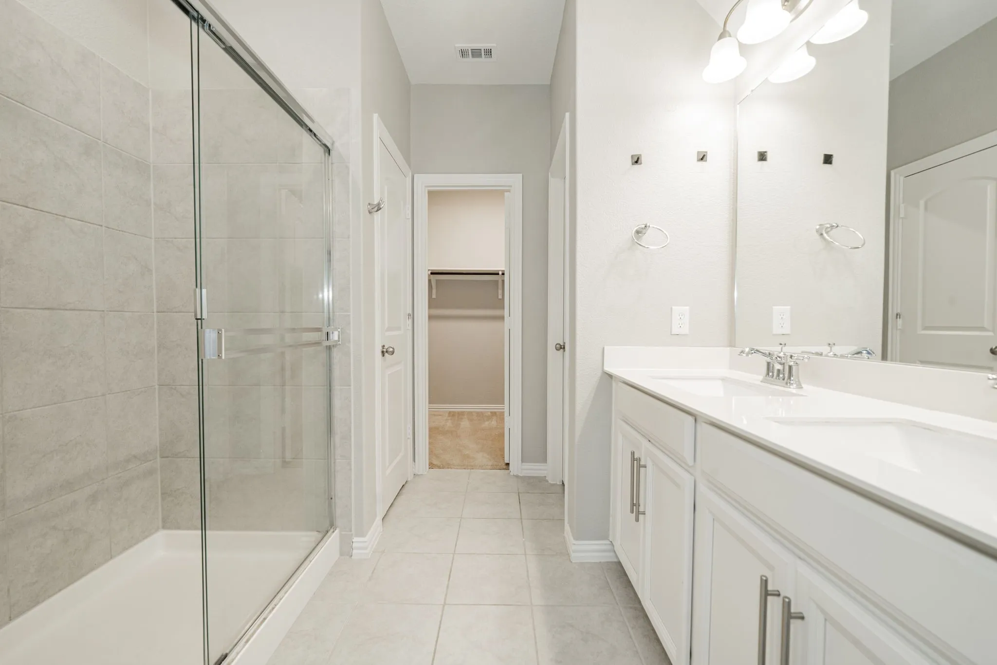 Dual vanity with white cabinetry and chrome hardware, glass-enclosed shower with tile surround, and tile flooring