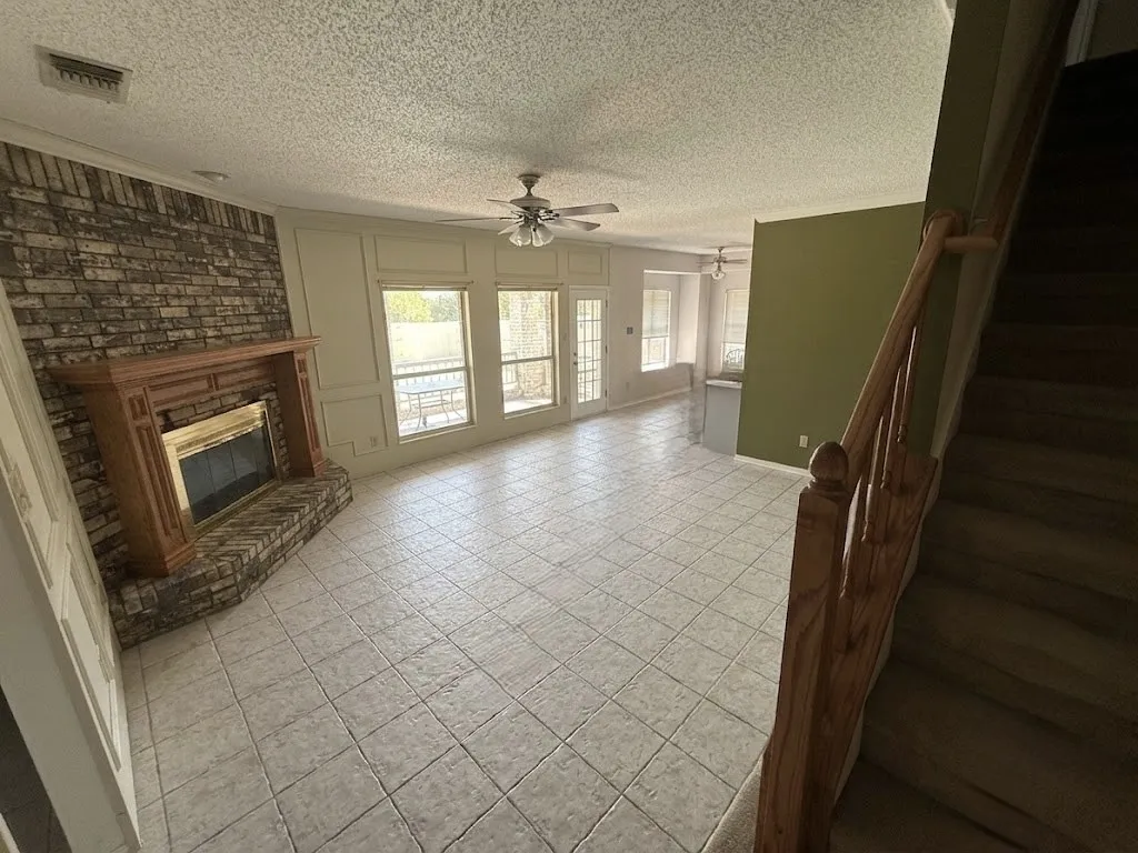 Spacious living area featuring tiled flooring, a brick fireplace with a wood-finish mantel, multiple large windows, and a ceiling fan