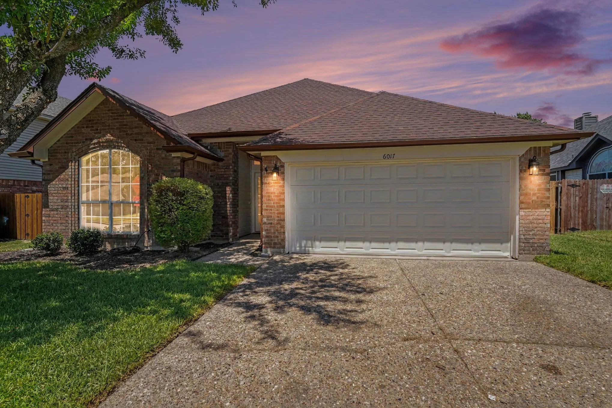 Brick exterior with an arched window, attached garage, concrete driveway, and mature landscaping