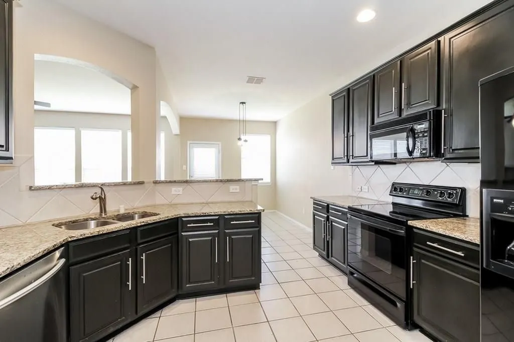 Kitchen featuring dark wood cabinetry, light-toned countertops, a stainless steel double basin sink, white tile flooring, and integrated black appliances