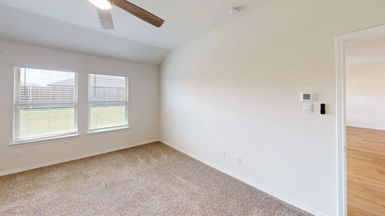 Carpeted flooring, vaulted ceiling, dual windows with blinds, ceiling fan with integrated lighting, and a wall-mounted thermostat