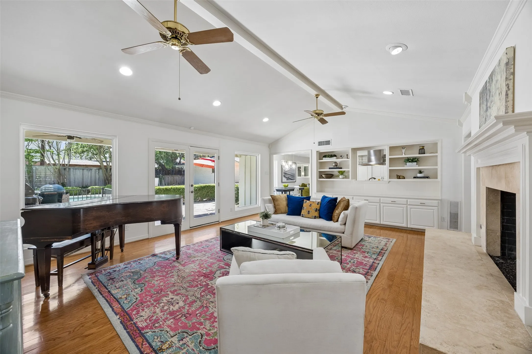 A wider view of the living room showcases the vaulted ceiling, fireplace, custom built-ins, wood floors, and seamless connection to the kitchen, backyard and pool.