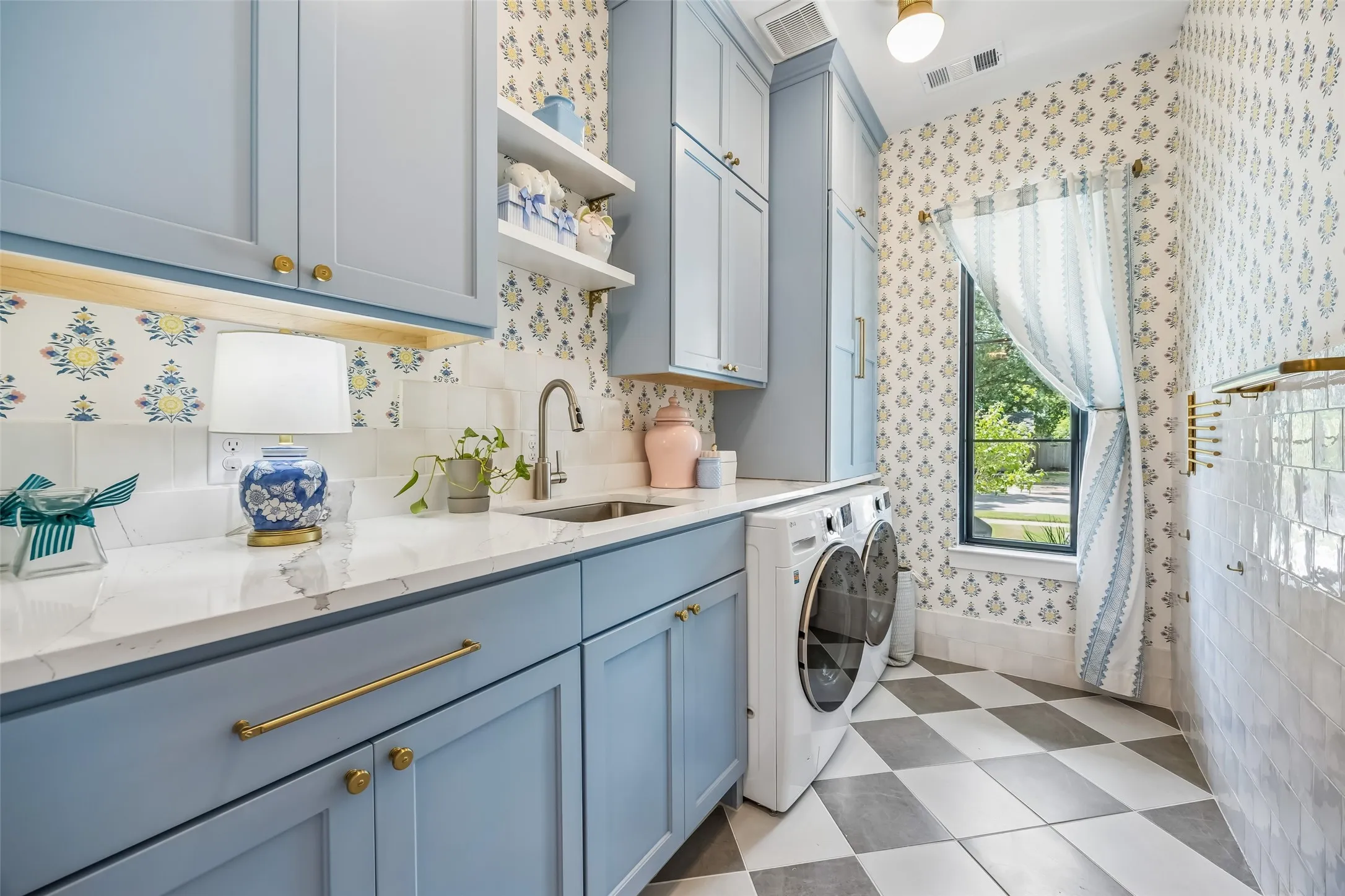 A beautifully designed laundry room combines function and style with custom blue cabinetry, generous storage, a utility sink, designer wallcovering and playful patterned flooring.