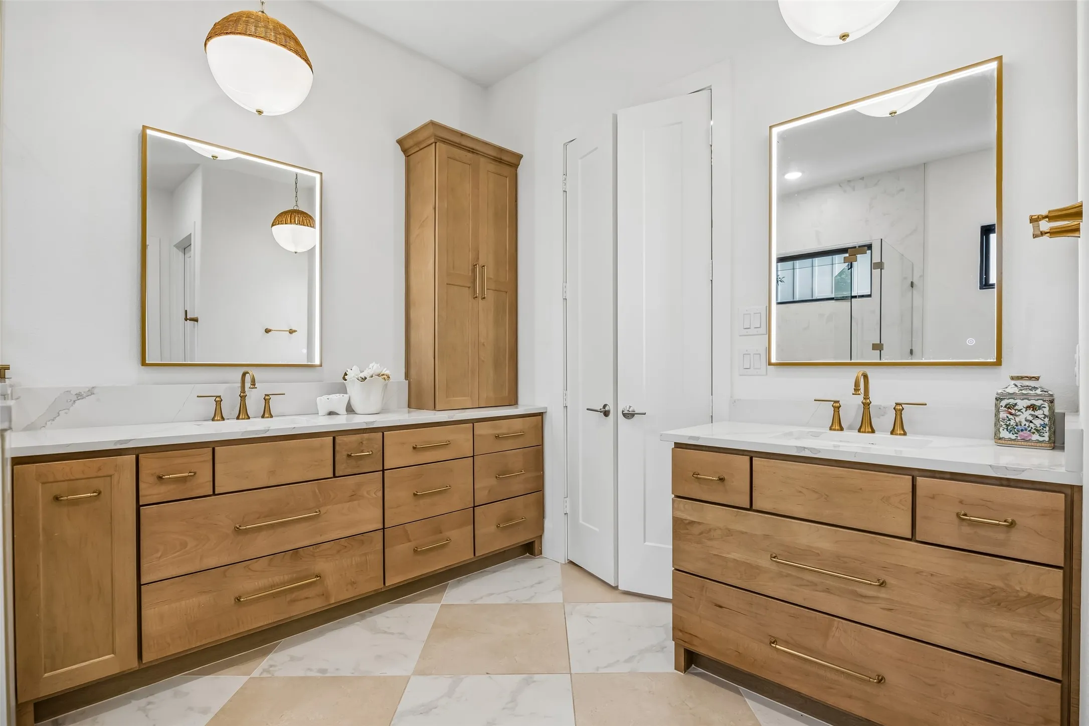 The primary bath features dual custom vanities with abundant storage, warm wood cabinetry, brass fixtures, illuminated mirrors and beautifully coordinated designer finishes.