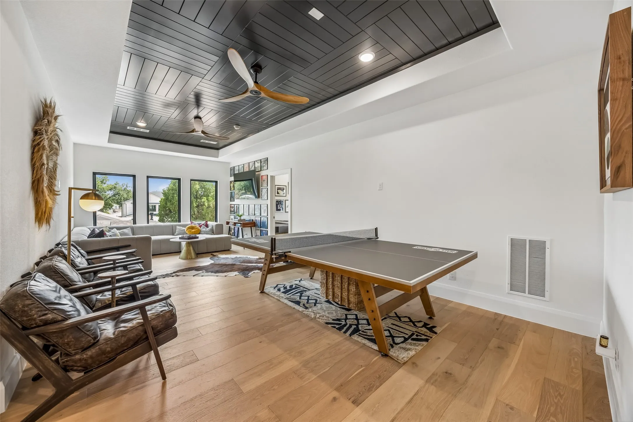 A spacious upstairs game room features wood floors, a dramatic paneled ceiling with dual fans and abundant natural light, creating a versatile space for relaxing or entertaining.