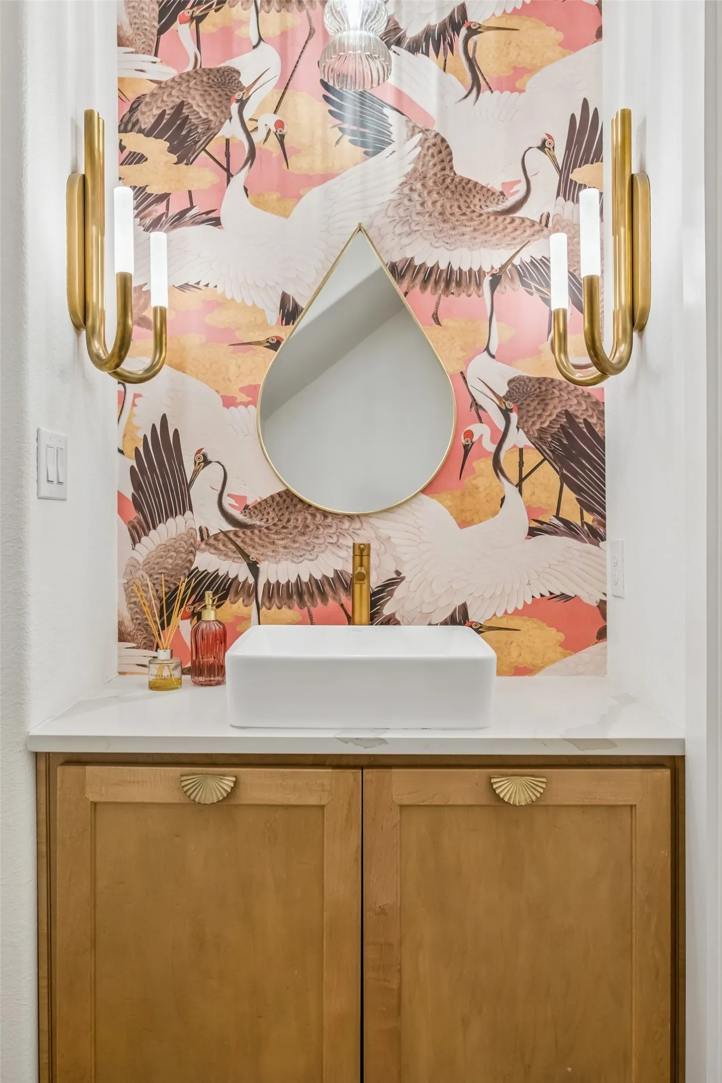 A jewel-box powder bath makes a memorable statement with whimsical designer wallcovering, sculptural brass sconces, warm wood cabinetry and distinctive custom details.