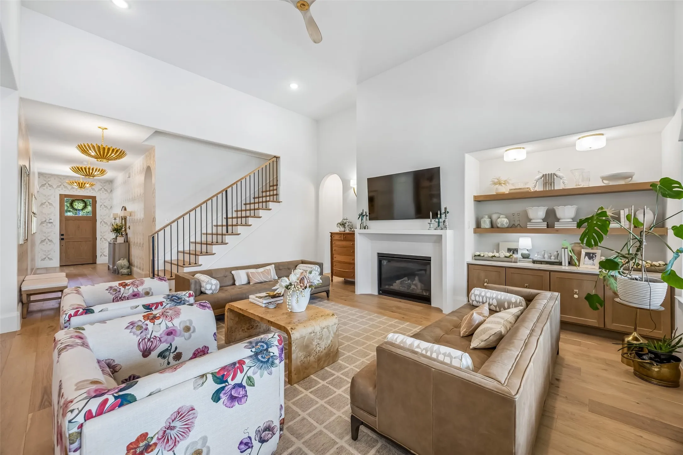 A dramatic living room features soaring ceilings, a sleek fireplace, custom built-ins with open shelving, wood floors and distinctive designer lighting that carries through the entry.