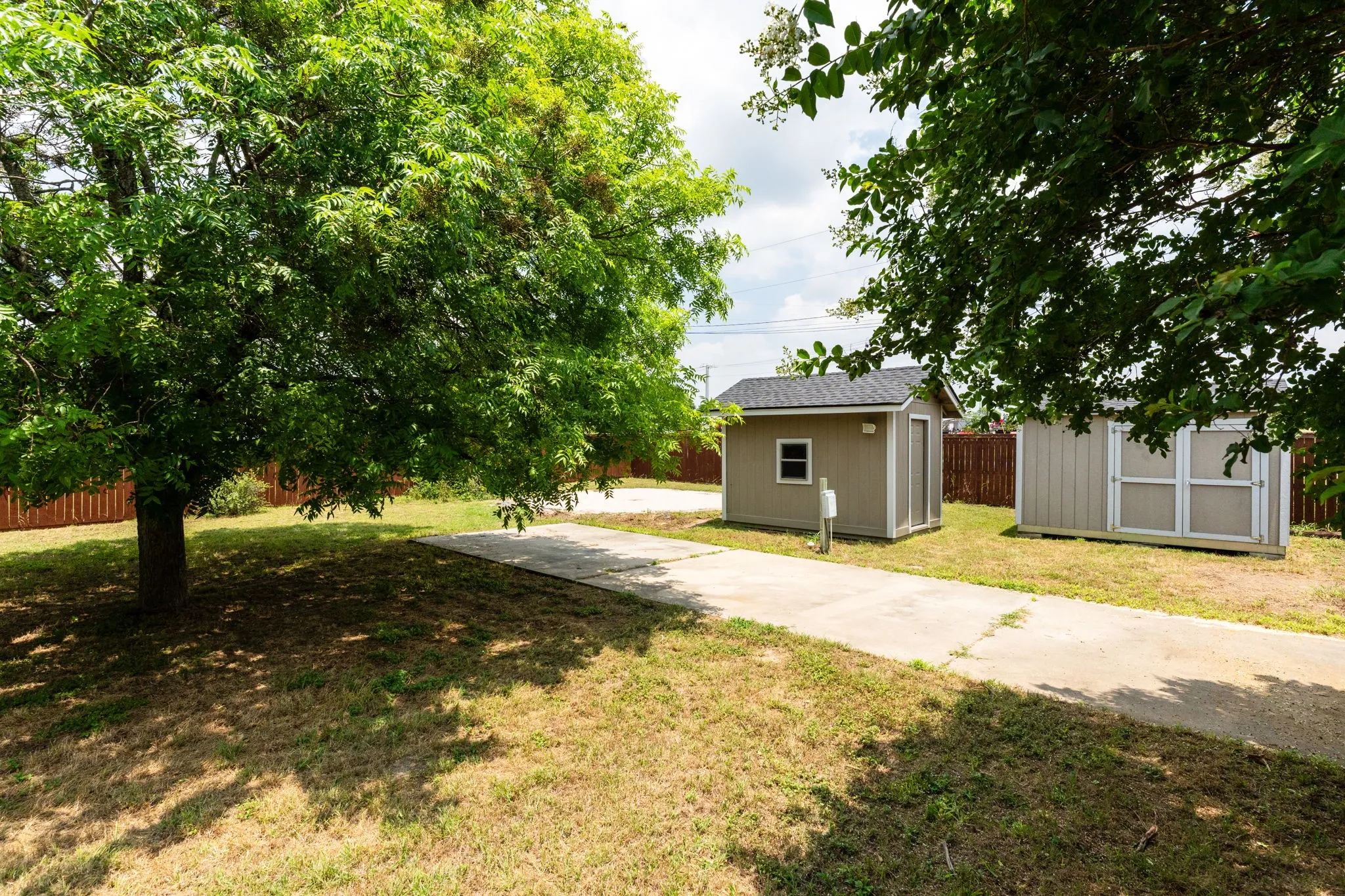 Expansive yard featuring mature trees, a concrete pathway, a privacy fence, and two storage sheds