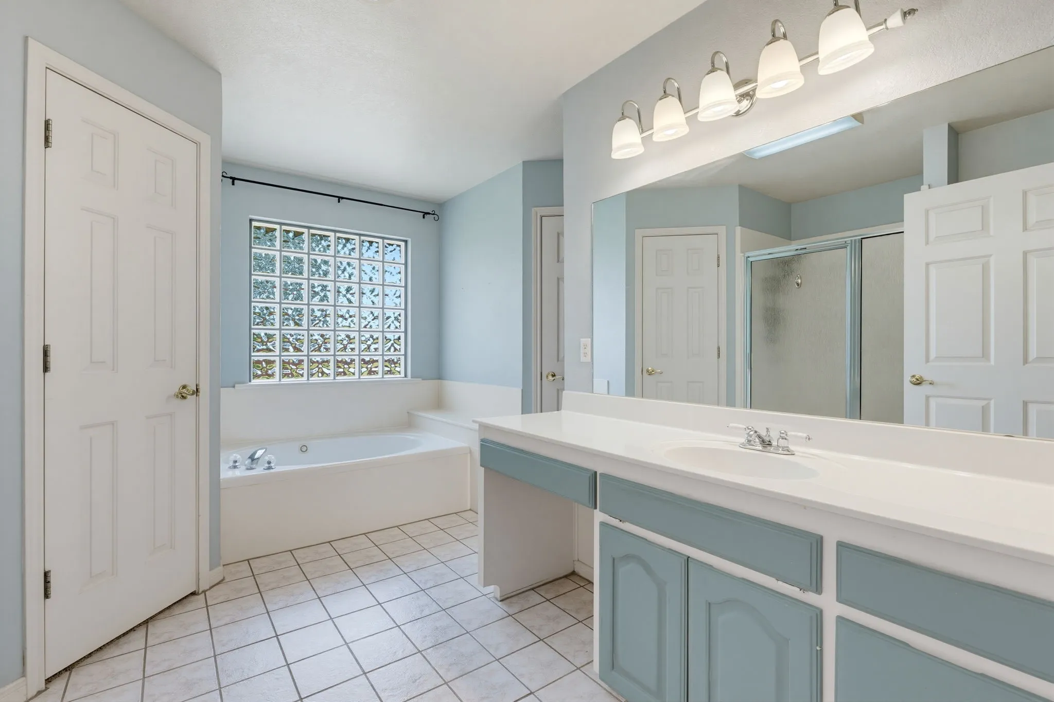 Dual vanity with undermount sink, large wall-mounted mirror, white ceramic tile flooring, built-in soaking tub, and glass block window