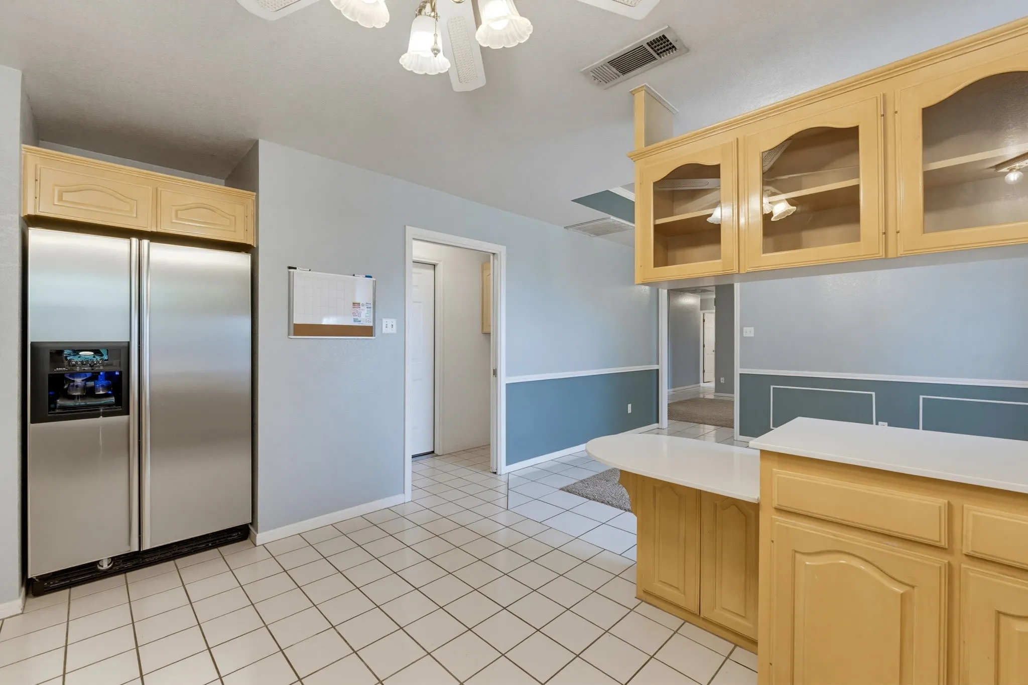 White tile flooring, stainless steel refrigerator, light wood-finish cabinetry, white countertops, and a ceiling fan with integrated lighting