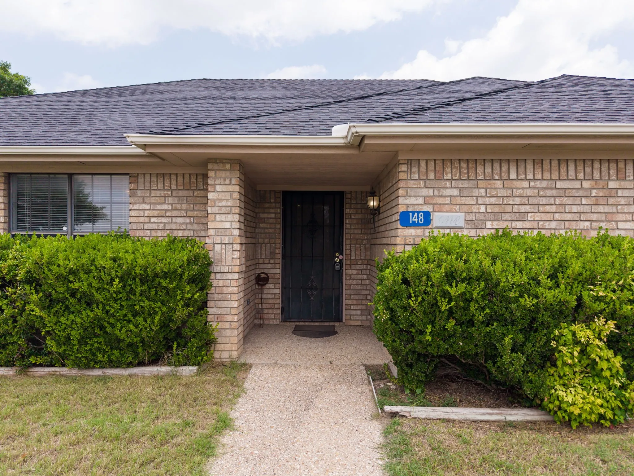 Brick exterior, dark shingle roof, concrete pathway, security screen door, and exterior lighting