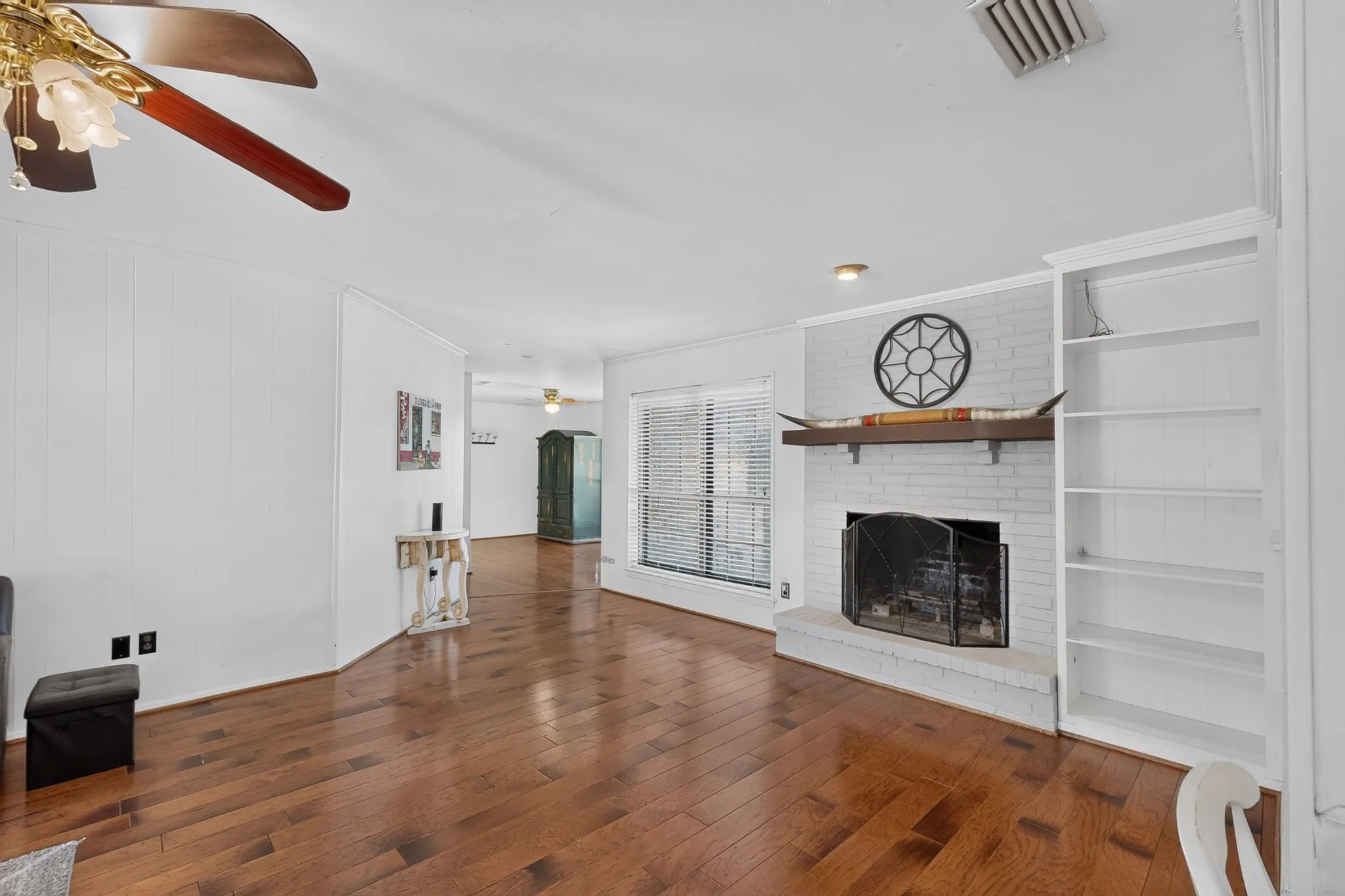 Wood-finish flooring, white brick fireplace, built-in shelving, white paneled walls, and a ceiling fan with light fixture