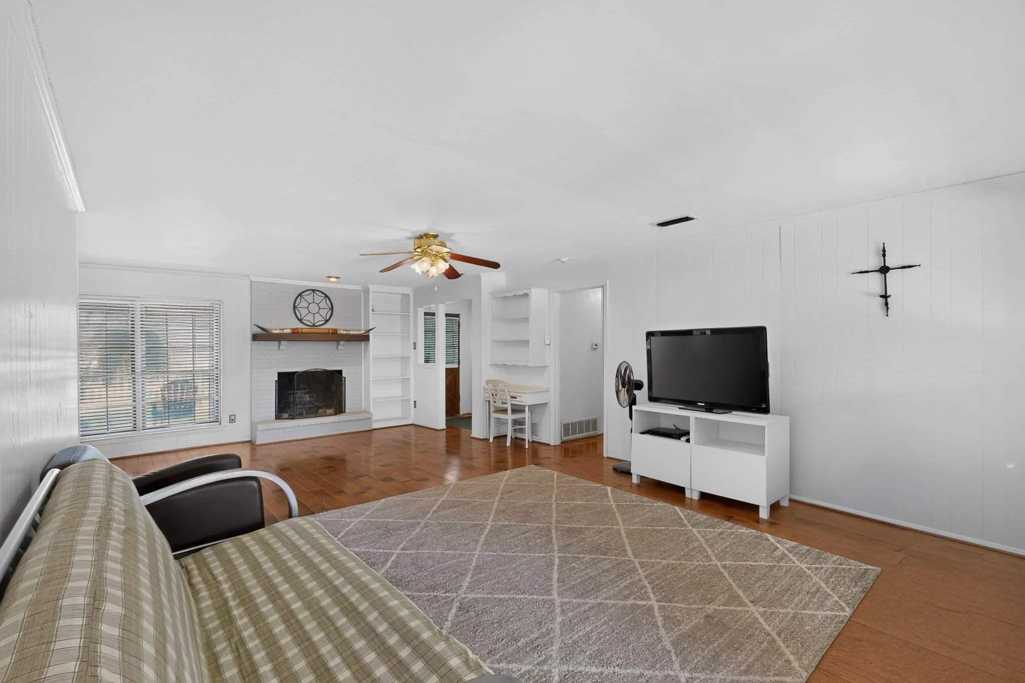 Wood-finish flooring throughout, white painted brick fireplace with hearth, built-in shelving, ceiling fan with integrated lighting, and window with blinds