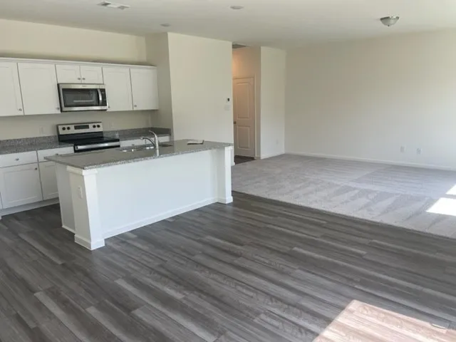 Open-concept living space with wood-finish flooring, white cabinetry, stainless steel appliances, and a kitchen island with stone-finish countertops