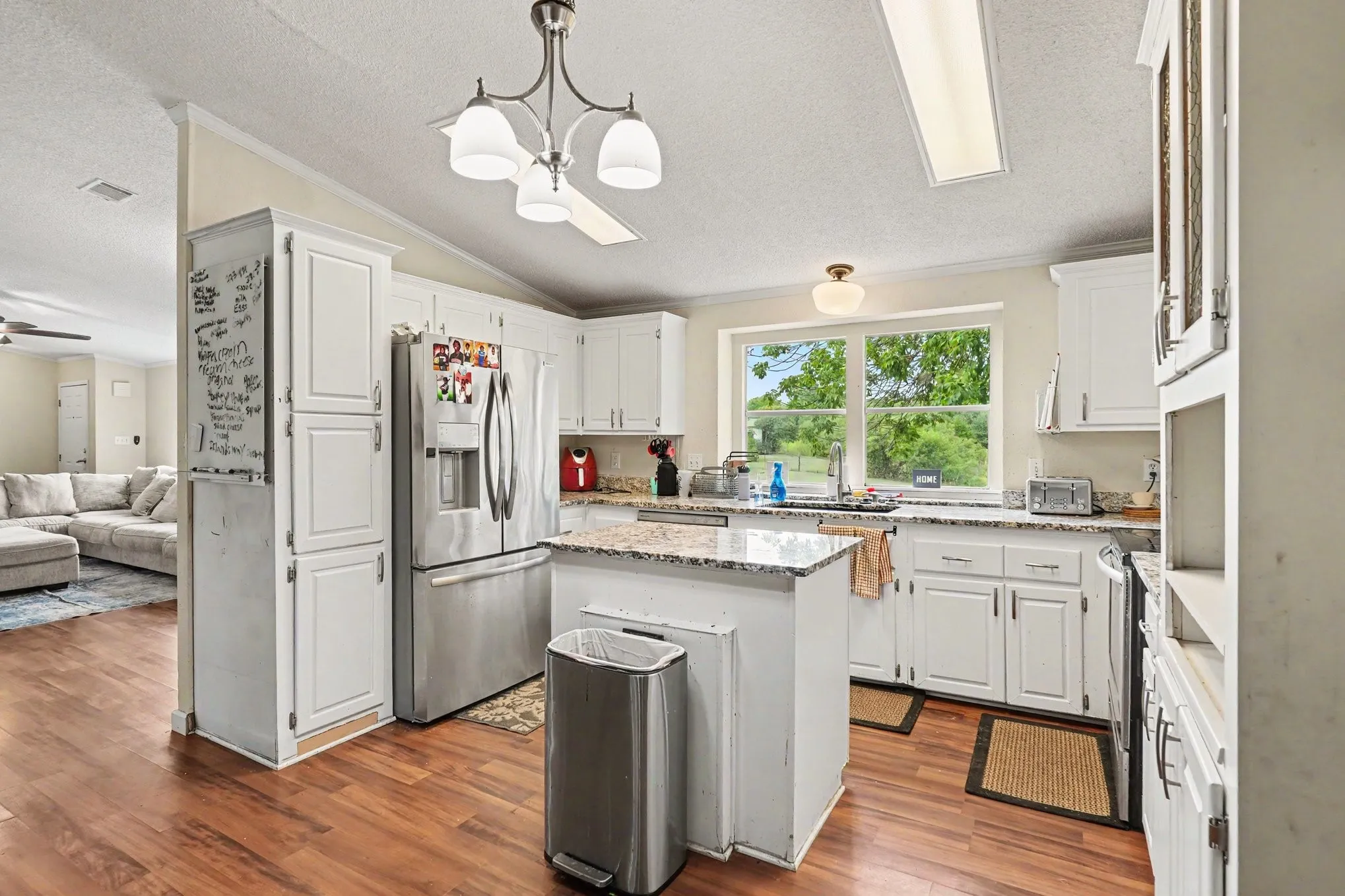 Bright kitchen featuring white cabinetry, a central island with a granite-finish countertop, stainless steel refrigerator, wood-finish flooring, and a large window above the sink