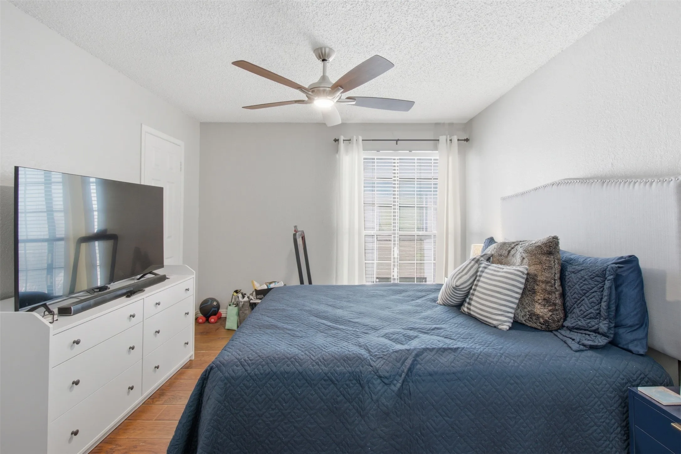 Bright secondary bedroom featuring hardwood flooring, a ceiling fan with integrated lighting, white built-in dresser with ample storage, and large window with plantation shutters allowing natural light throughout the space
