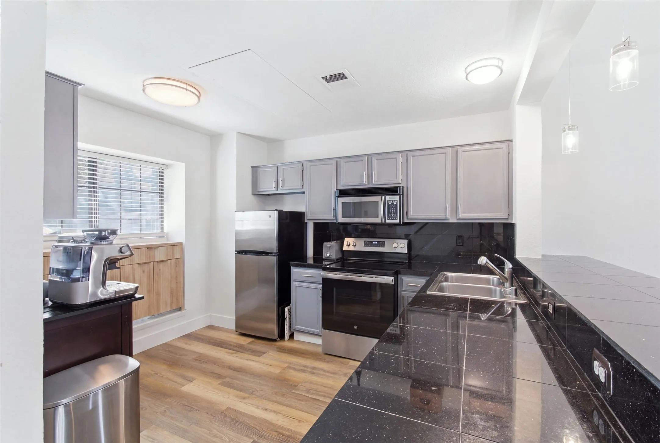 Modern kitchen featuring stainless steel appliances, dark stone countertops with waterfall edge detail, gray cabinetry with ample storage, wood-finish flooring, and recessed ceiling lighting