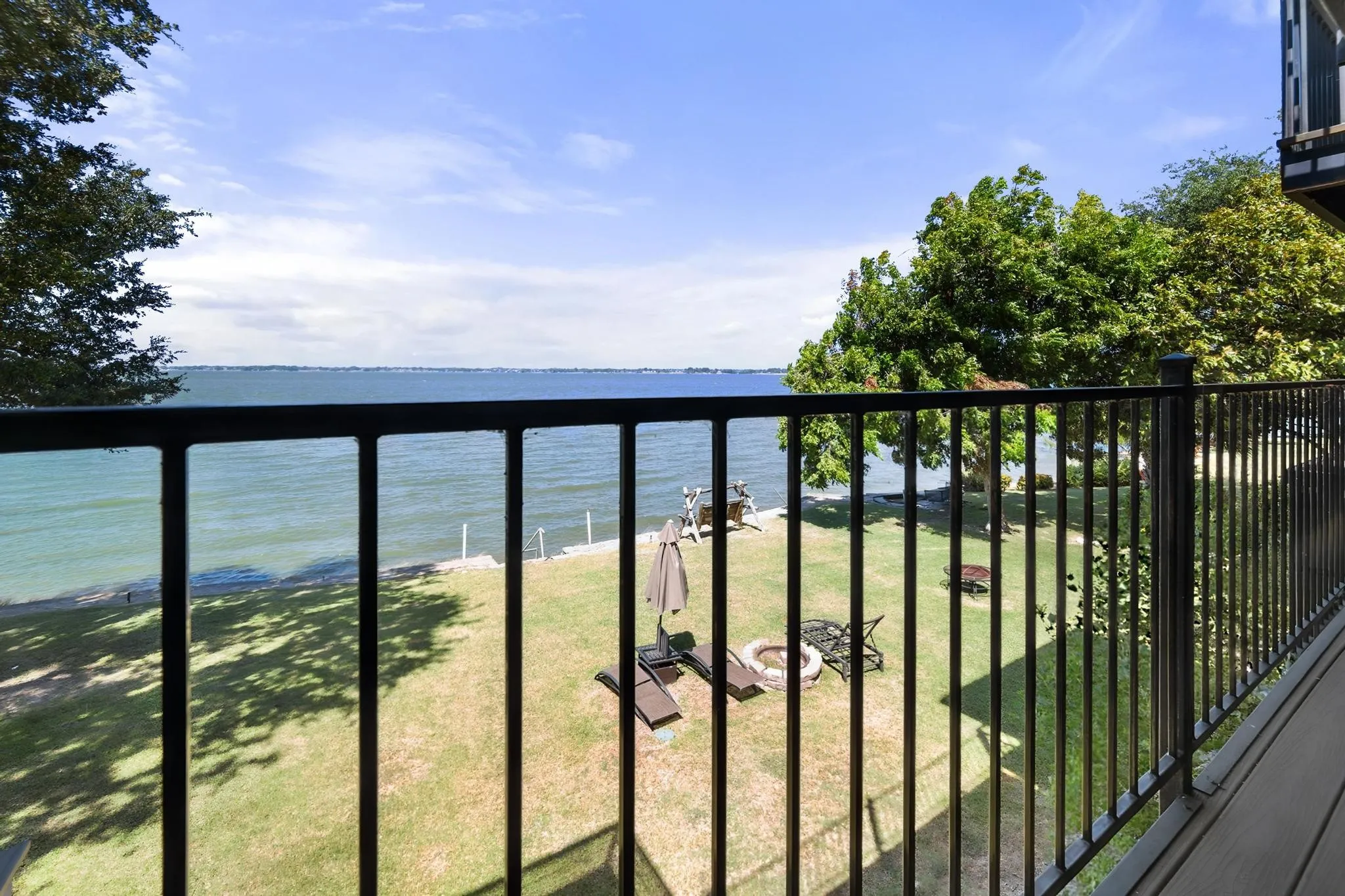 Balcony featuring black metal railings and wood-finish decking overlooking a body of water, a grassy yard, and mature trees