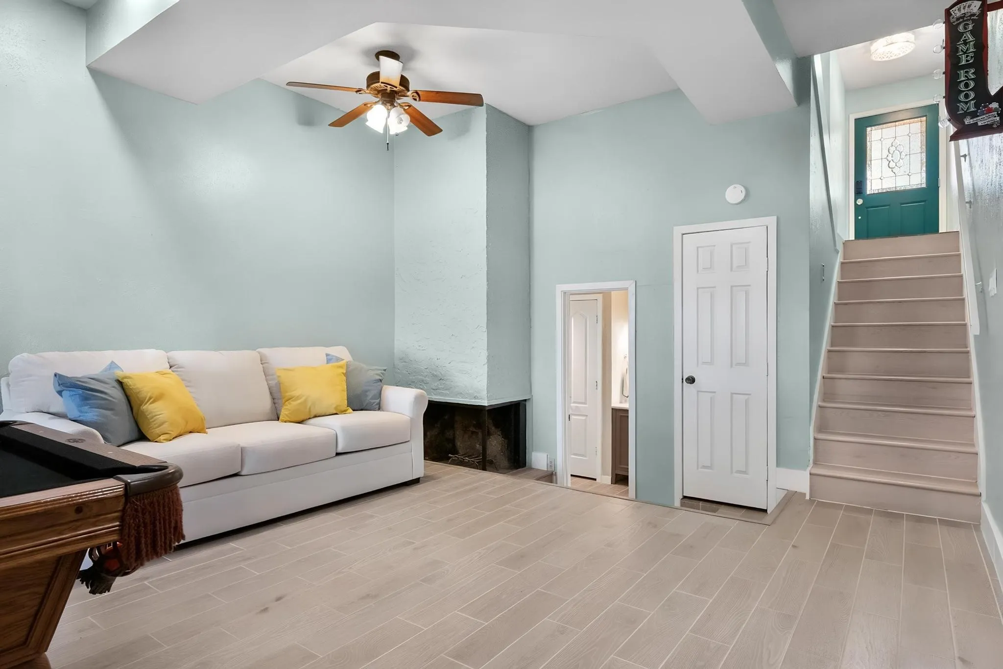 Wood-finish flooring throughout, featuring a ceiling fan, a built-in fireplace structure, and a staircase with a landing leading to a paneled door with leaded glass