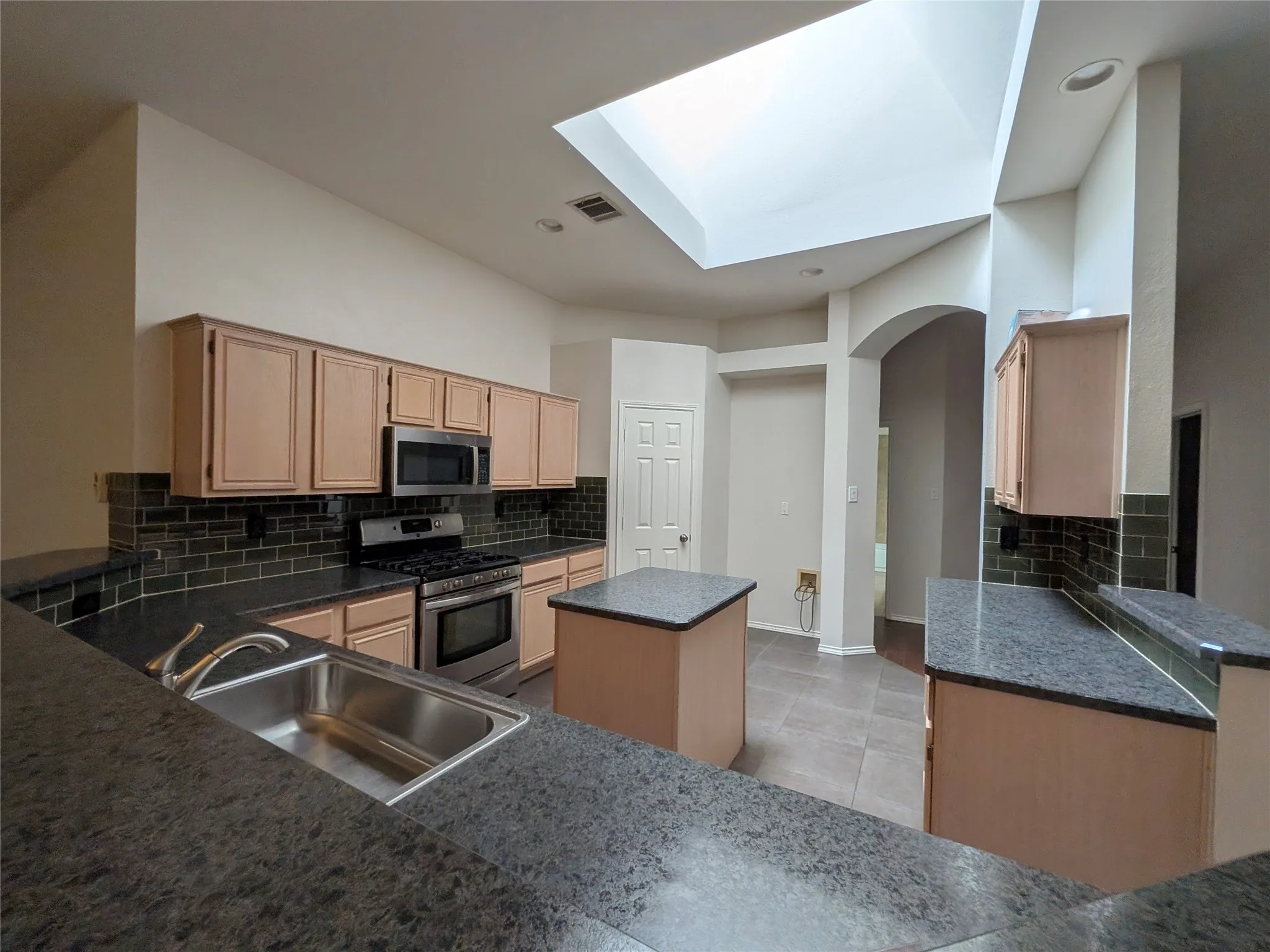 Kitchen featuring an integrated stainless steel sink, dark granite-look countertops, subway tile backsplash, and light wood-finish cabinetry
