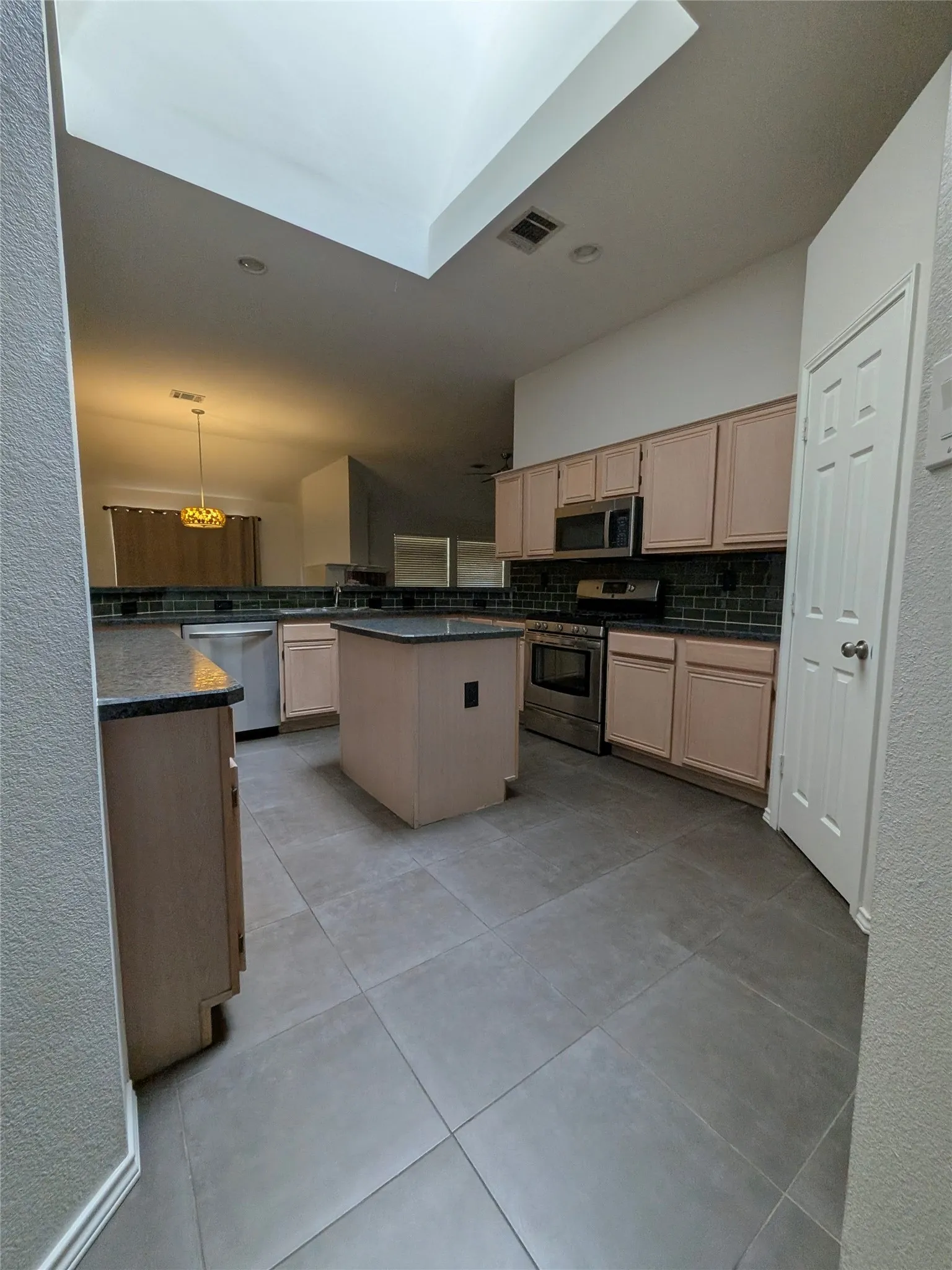 Skylight feature with recessed lighting, central kitchen island, gray tile flooring, light wood-finish cabinetry, and stainless steel appliances