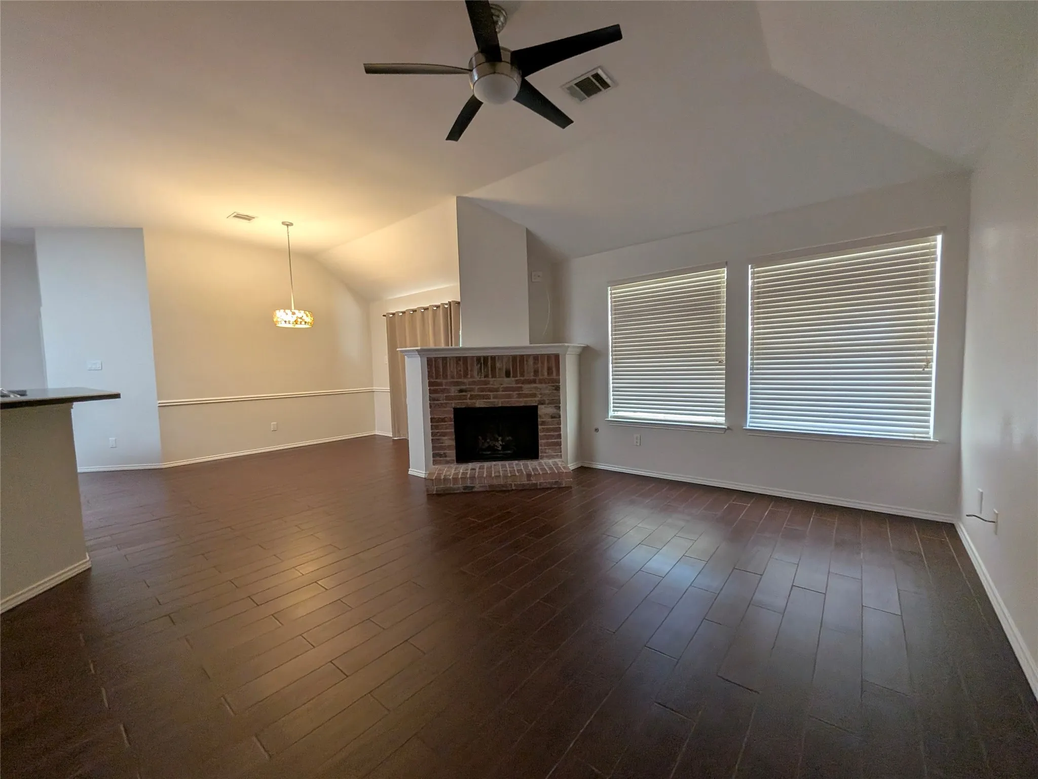 Wood-finish flooring, brick fireplace with mantel, ceiling fan, and multiple windows with blinds