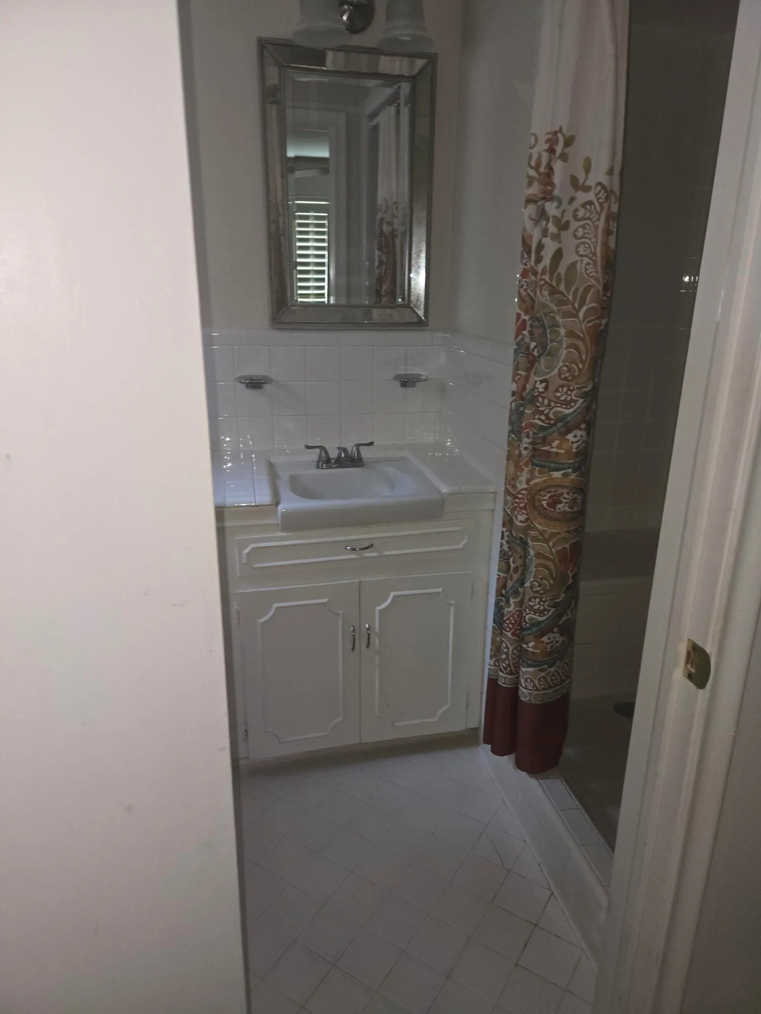 White tile flooring, white vanity with paneled doors, square undermount sink, mirrored vanity light fixture, and tiled shower surround