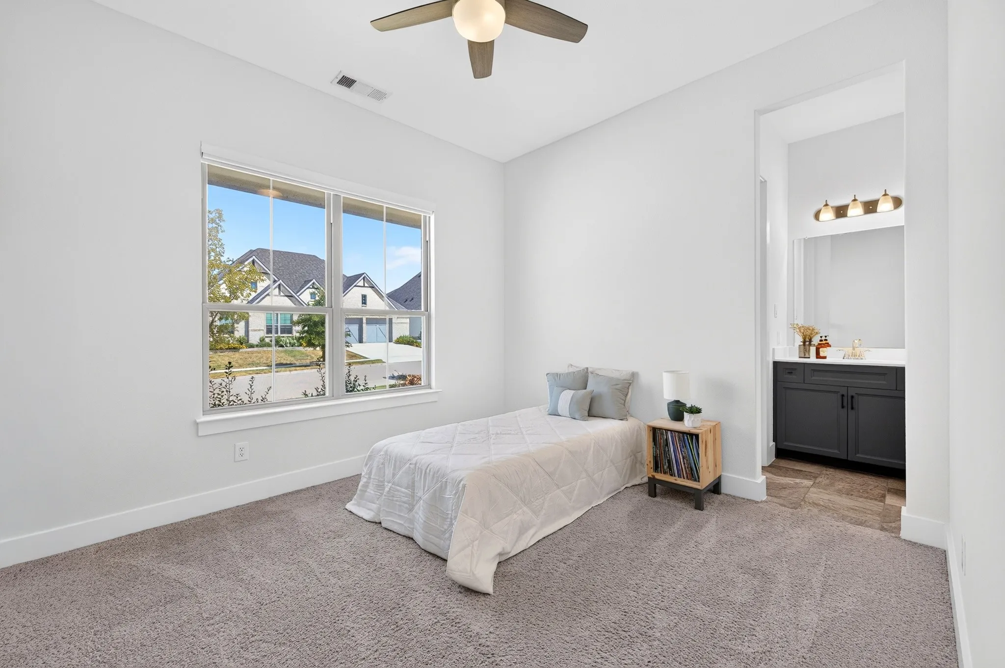 Plush carpeted room featuring a large window, a ceiling fan, and access to a bathroom with a dark vanity, white countertop, and three-light fixture