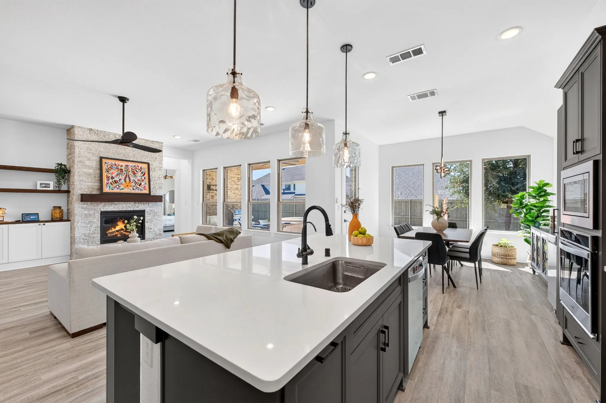 Kitchen island with stone countertop and undermount sink, pendant lighting, wood-finish flooring, stone-clad fireplace with wood mantel, and built-in white cabinetry with shelving