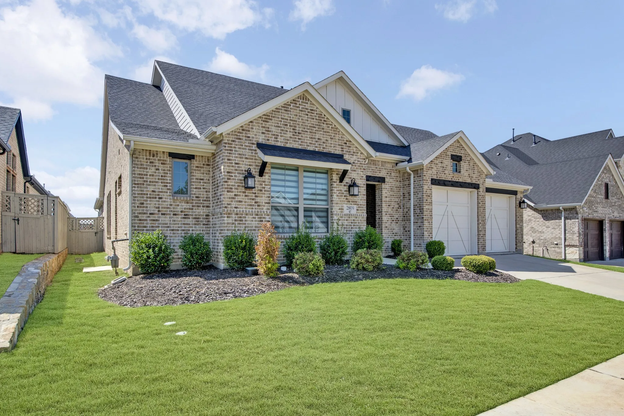 Brick exterior with a dark shingle roof, two-car garage with carriage-style doors, landscaped front yard, and a paved driveway