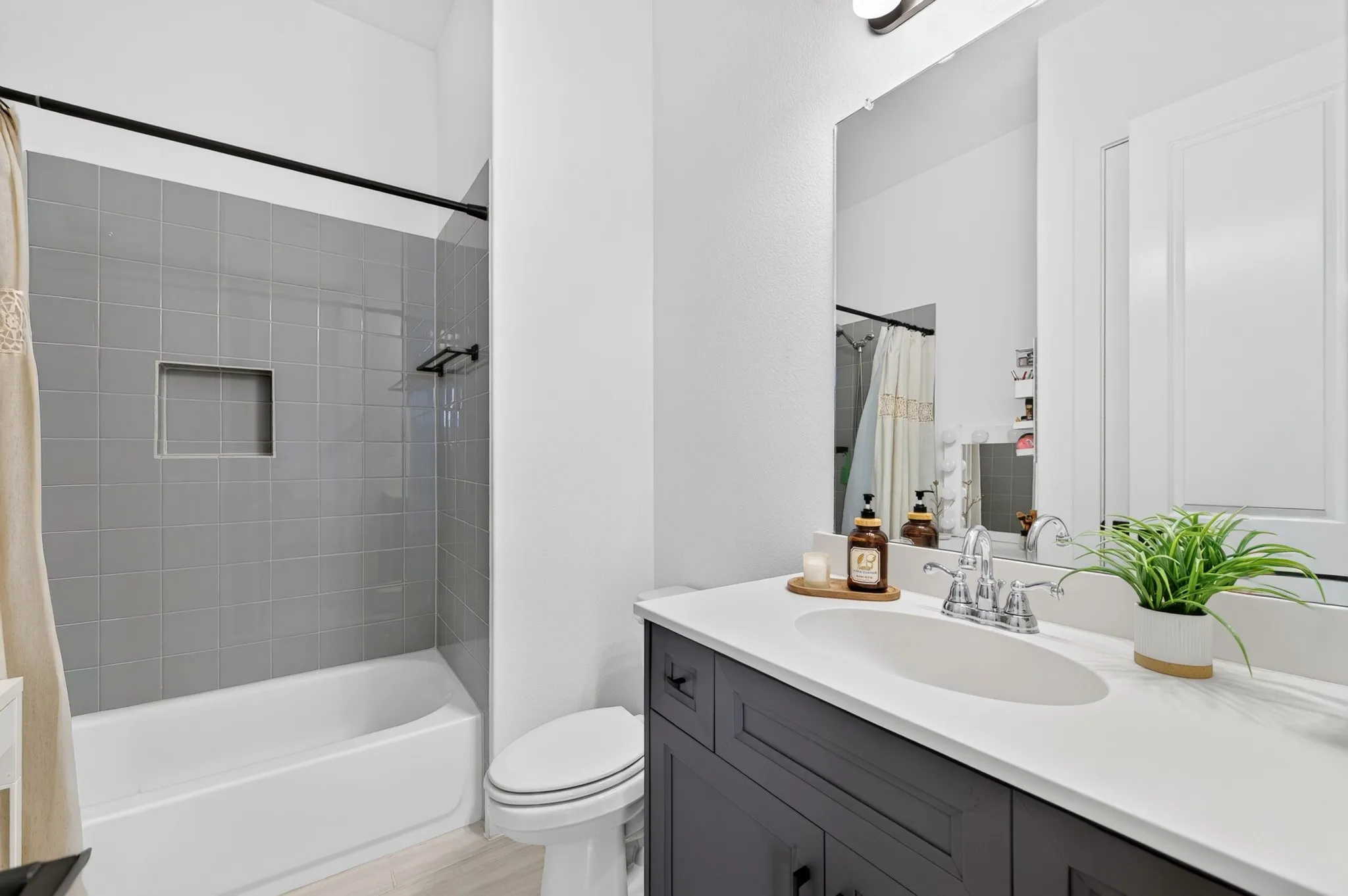Bathtub with gray tile surround and recessed niche, dark cabinetry with chrome hardware, integrated sink with chrome faucet, wall-mounted mirror, and wood-finish flooring