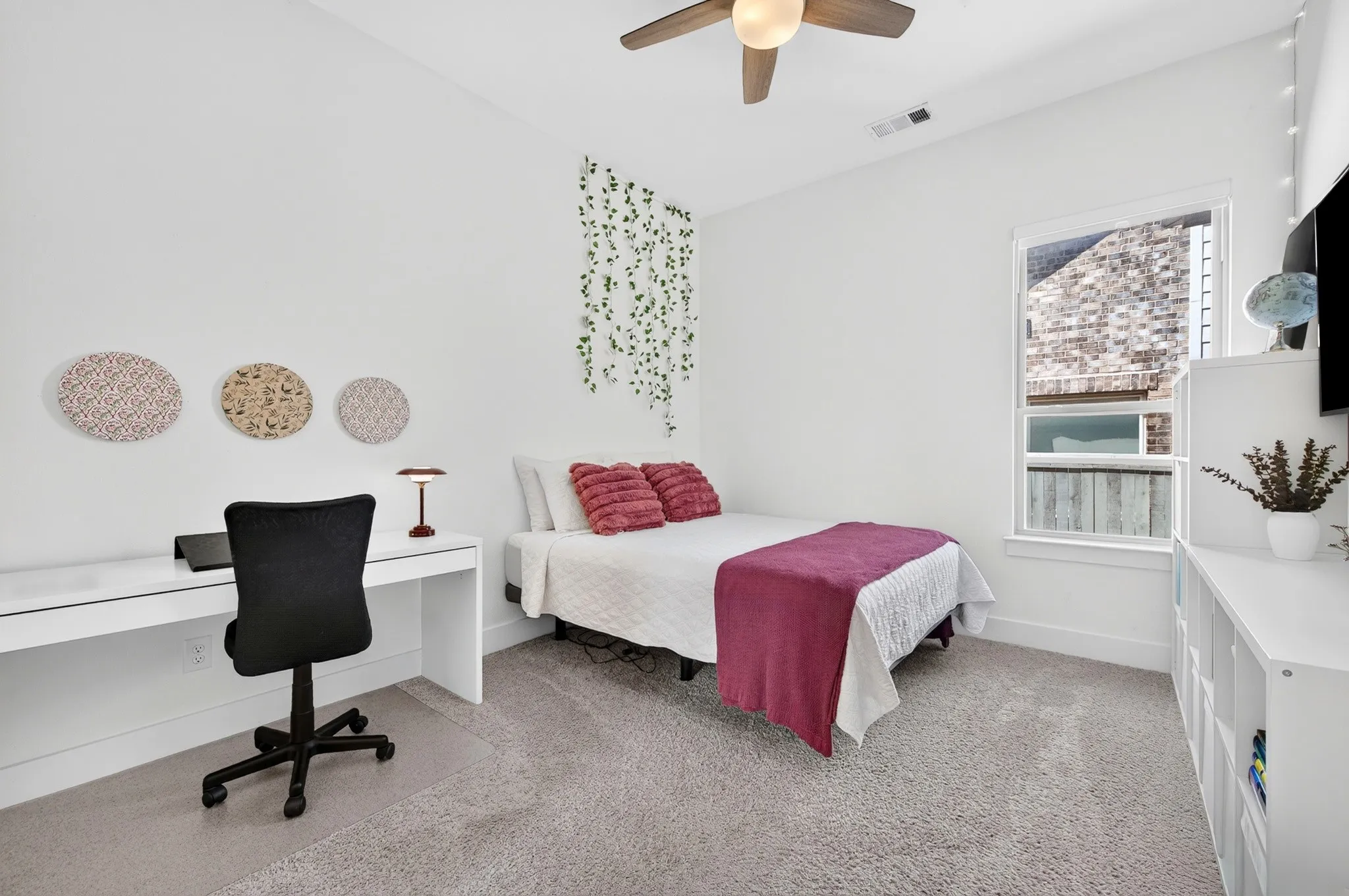 Plush carpet flooring, a built-in white desk, white walls, a multi-pane window, and a ceiling fan with integrated lighting
