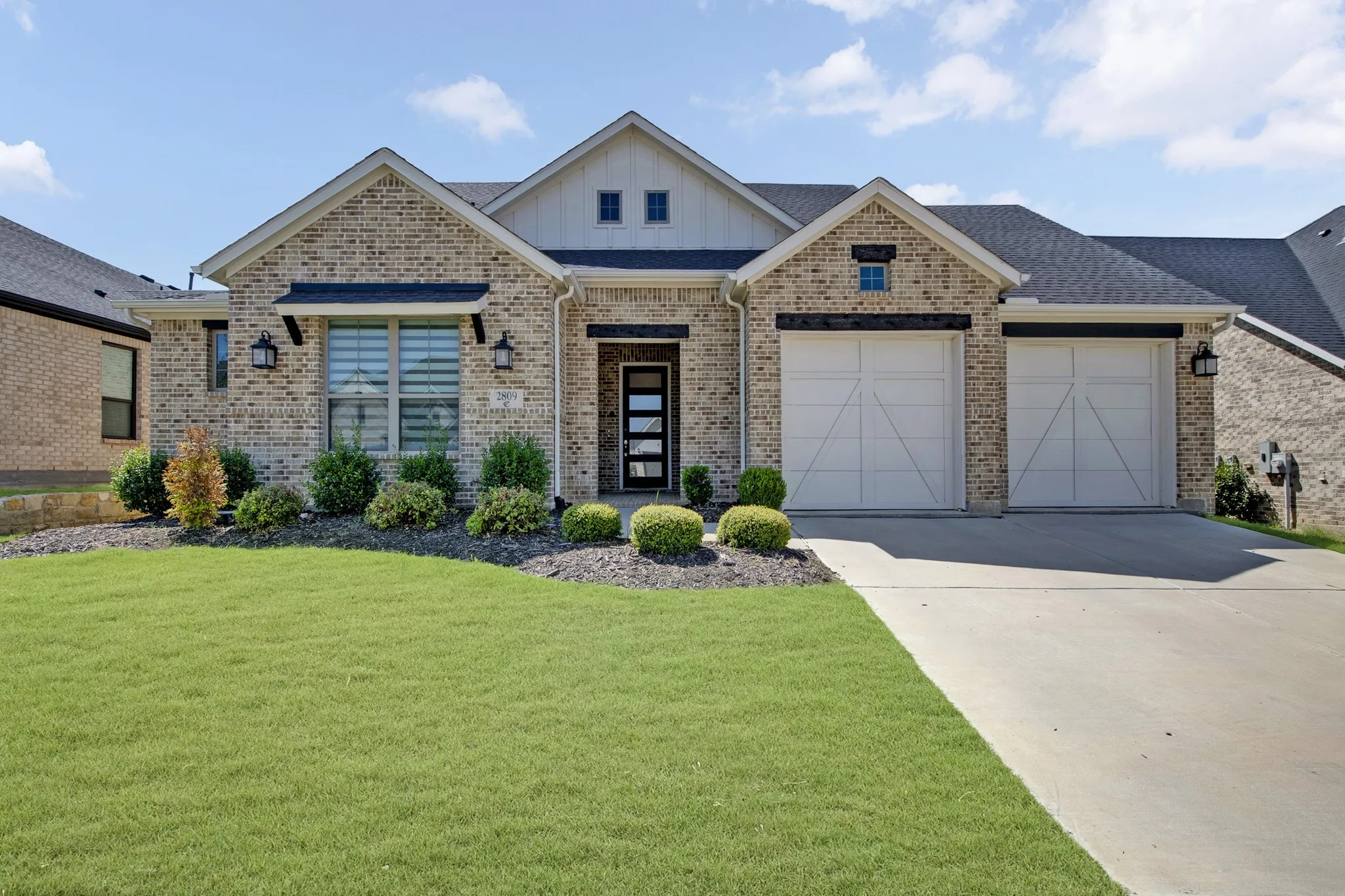 Brick exterior with board-and-batten gables, a two-car garage with carriage-style doors, a concrete driveway, and mature landscaping