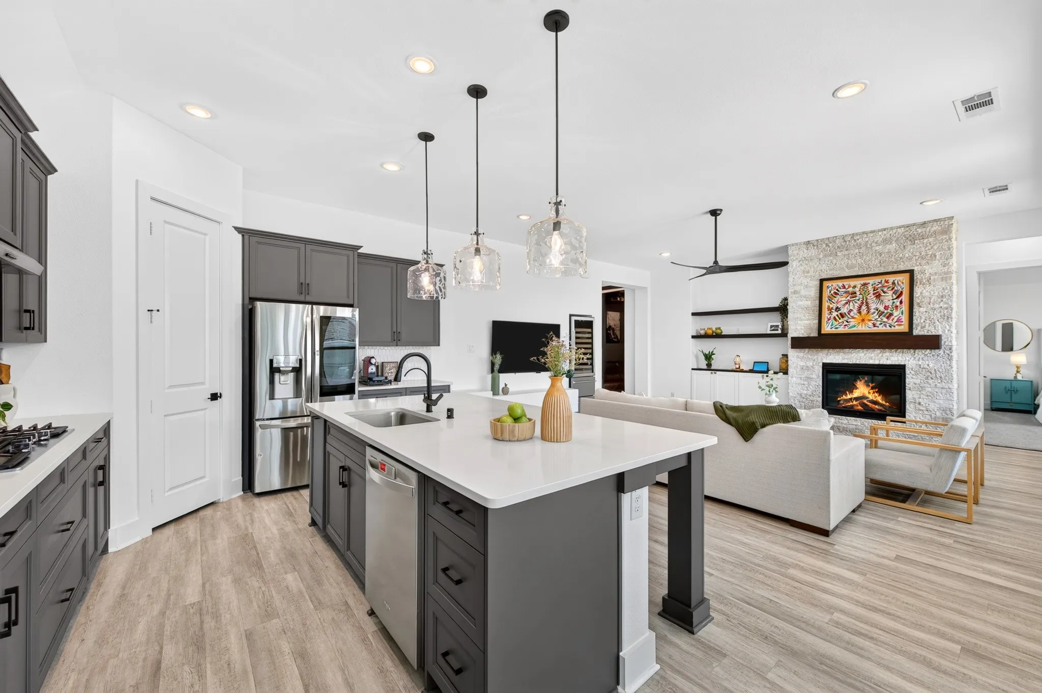 Open-concept kitchen and living area featuring a stone fireplace, wood-finish flooring, recessed lighting, a kitchen island with pendant lighting, and stainless steel appliances