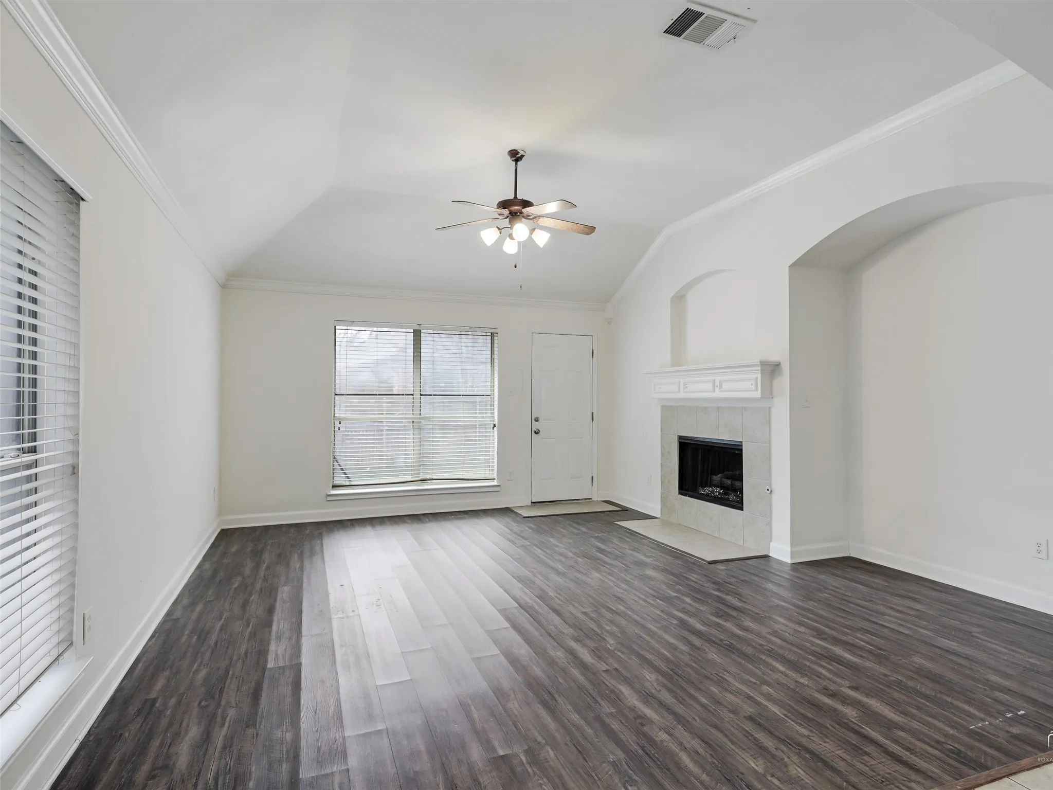 Vaulted ceiling with crown molding, wood-finish flooring, fireplace with mantel and tile surround, and an arched wall feature
