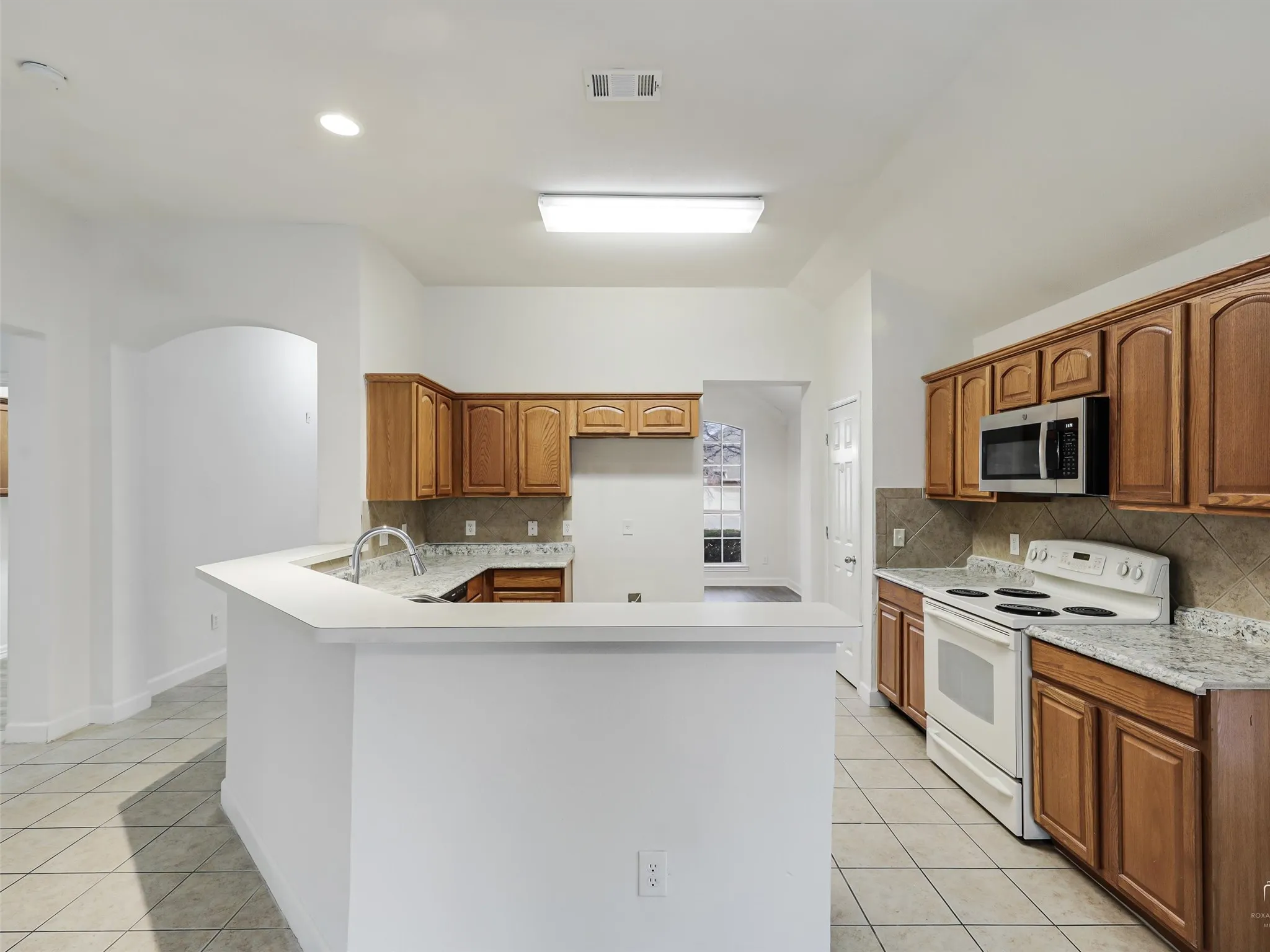Kitchen with wood cabinetry, granite-style countertops, tile backsplash, stainless steel microwave, and white electric range