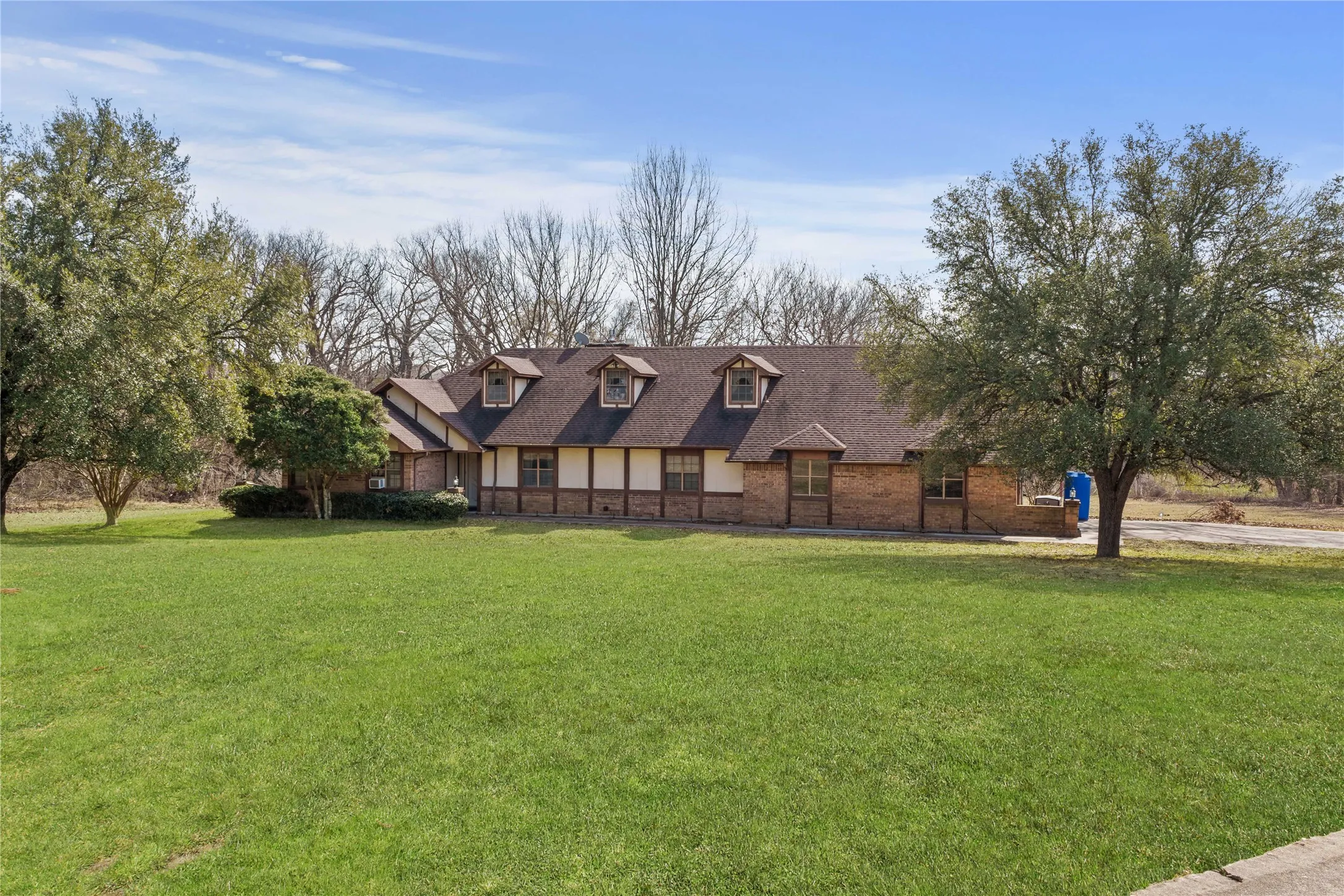 Brick and stucco exterior with dormer windows, featuring a prominent pitched roofline, surrounded by mature trees and a manicured lawn