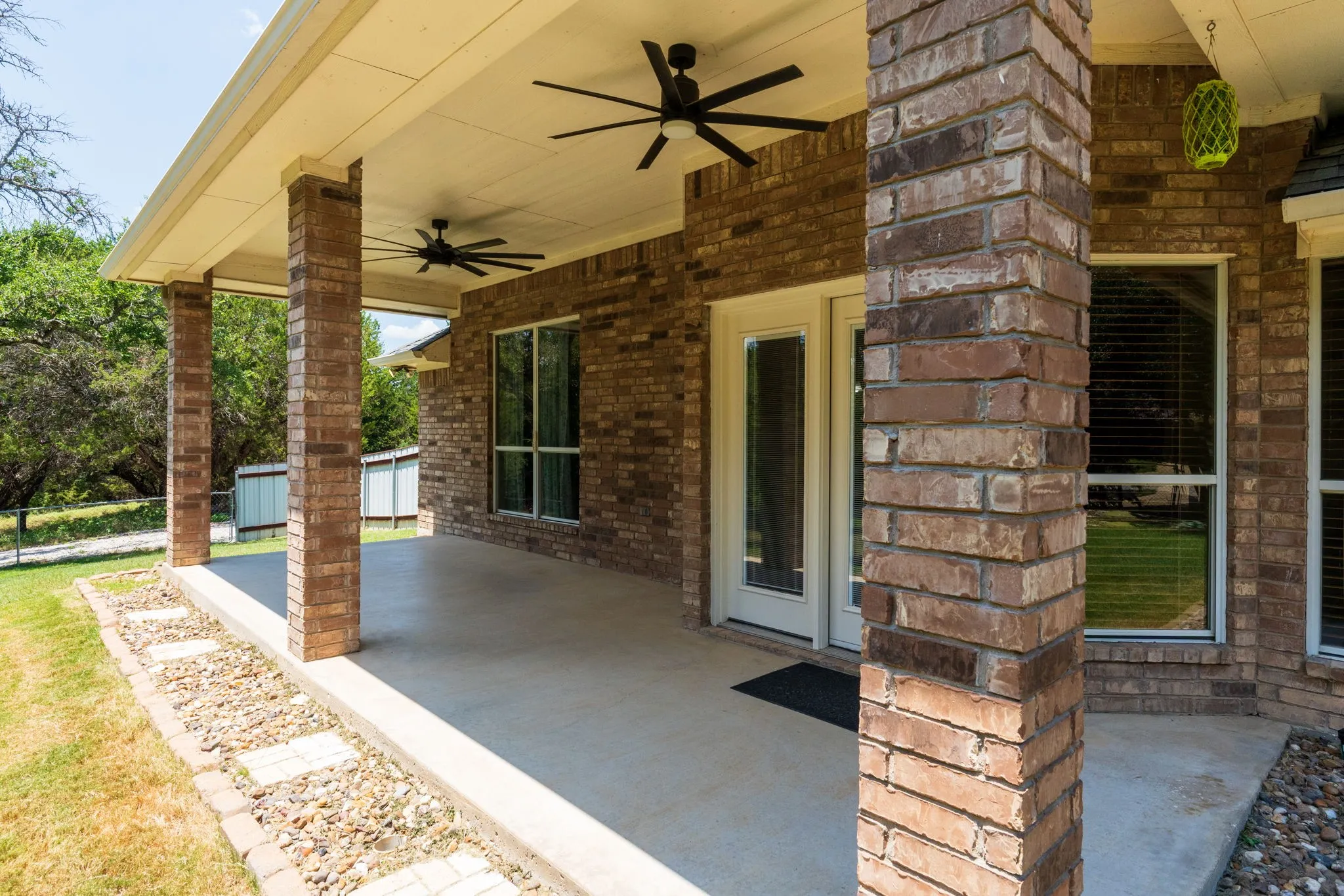 Covered patio featuring brick pillars, two ceiling fans, a concrete surface, and a stone-lined garden bed