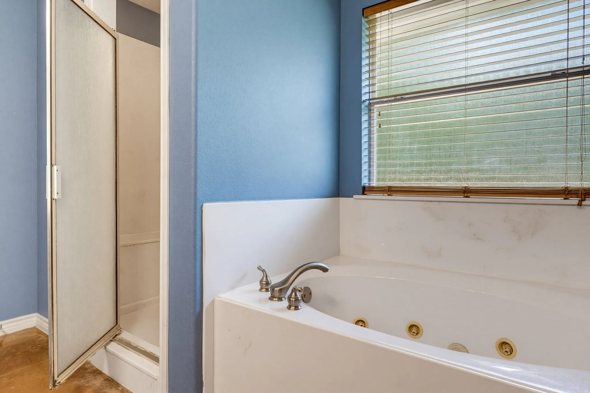 Jetted soaking tub with deck-mounted brushed nickel fixtures, an adjacent shower enclosure, and a window with blinds