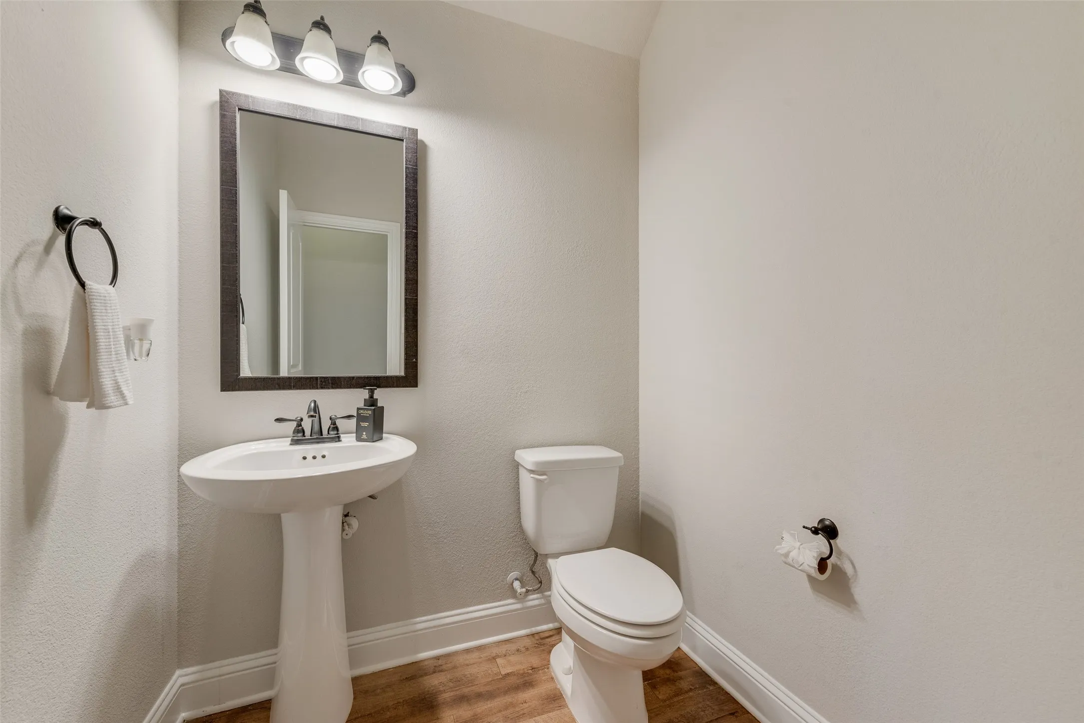 Pedestal sink with a dual-handle faucet, rectangular framed mirror, three-light vanity fixture, and wood-finish flooring
