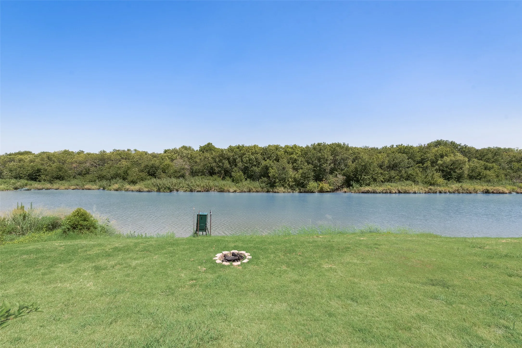 Fish feeder. Expansive grassy area with a stone-edged fire pit, bordering a serene body of water with a natural tree line